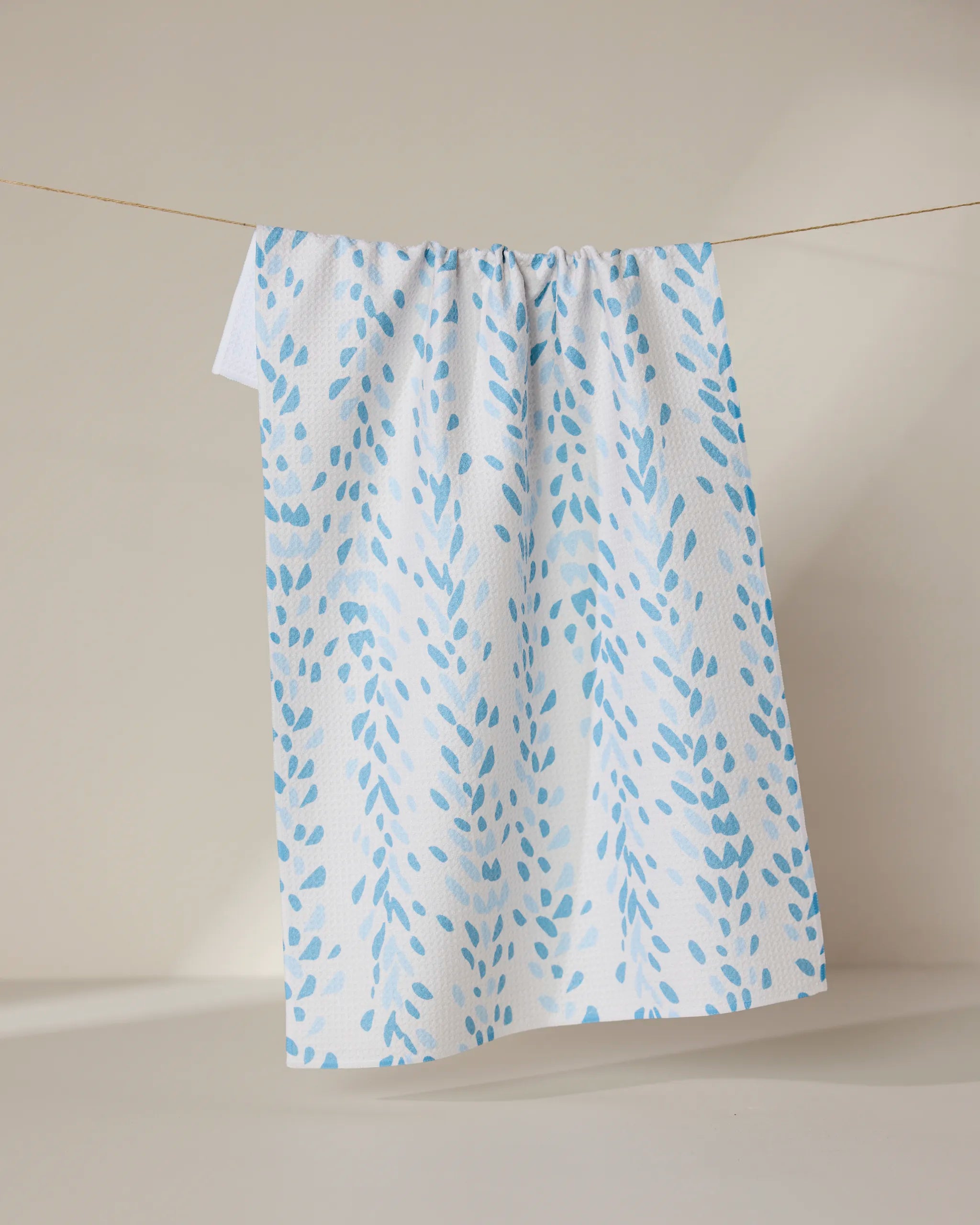 The GEOMETRY Reeds Printed - Midday is a white tea towel featuring light blue leaf-like patterns, crafted from recycled materials, shown hanging neatly on a clothesline against a neutral background.