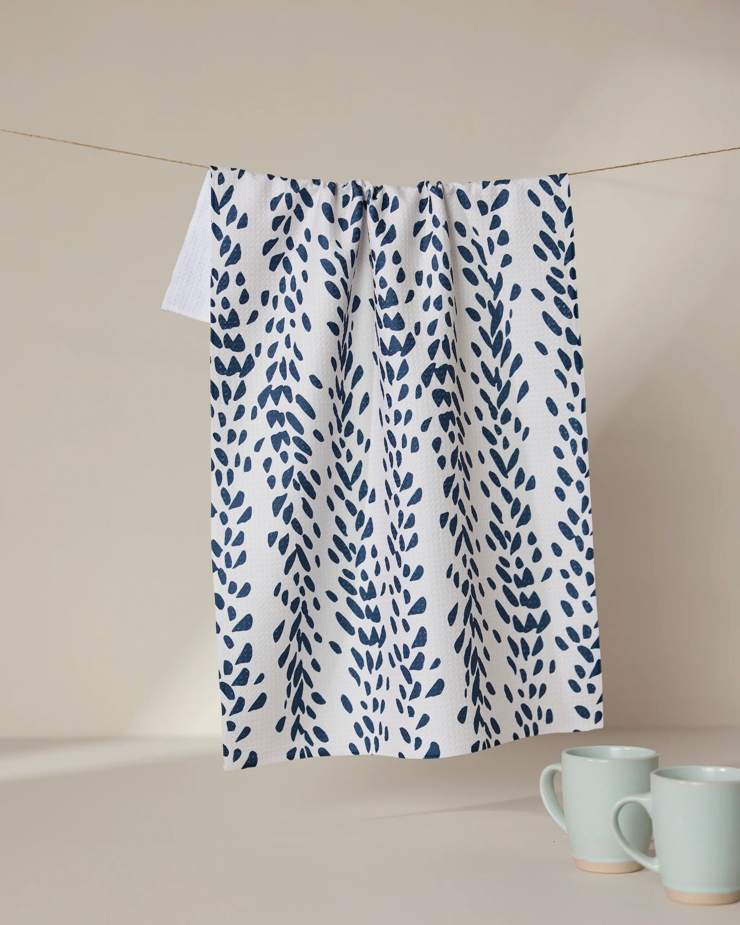 A GEOMETRY Reeds Printed - Midnight white kitchen tea towel with a blue leaf design hangs from a clothesline. Below, two light blue ceramic mugs rest on a white surface against a plain light beige background.