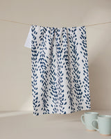 A GEOMETRY Reeds Printed - Midnight white kitchen tea towel with a blue leaf design hangs from a clothesline. Below, two light blue ceramic mugs rest on a white surface against a plain light beige background.