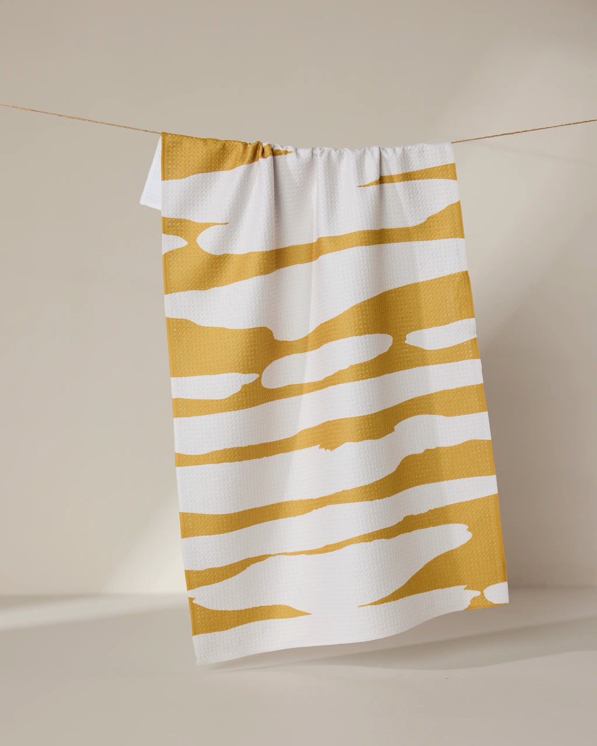 The Reflected - Midday tea towel by GEOMETRY, crafted from recycled materials, features irregular mustard-yellow horizontal stripes on white and hangs against a neutral beige background.