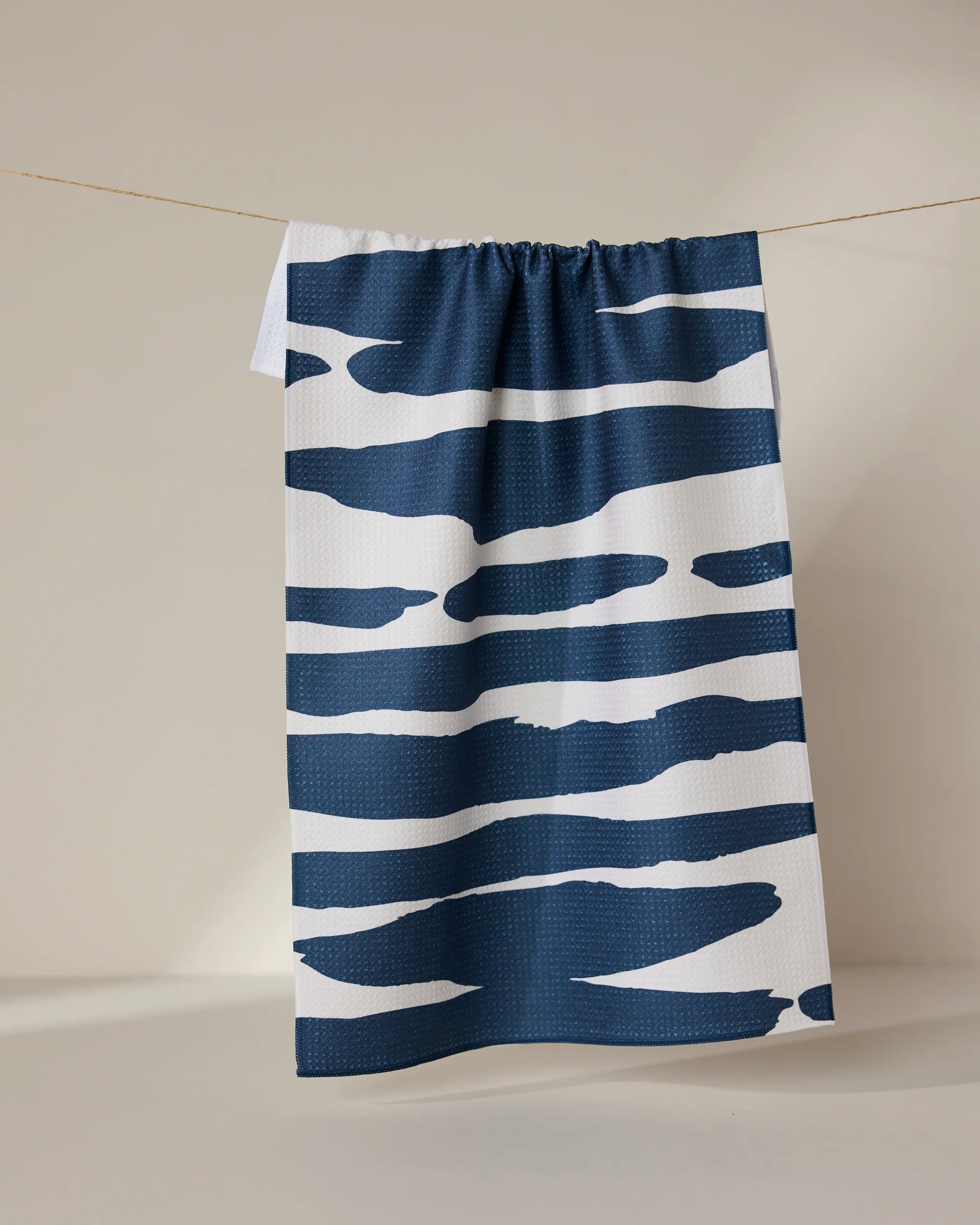The GEOMETRY Reflected - Midnight tea towel, crafted from ultra-absorbent recycled materials, features bold, irregular navy blue stripes on white and hangs on a thin clothesline against a neutral backdrop.