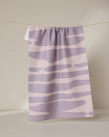 The GEOMETRY Reflected - Sunset tea towel, featuring pastel purple and cream abstract wavy stripes and made from recycled materials, hangs on a clothesline against a neutral background.
