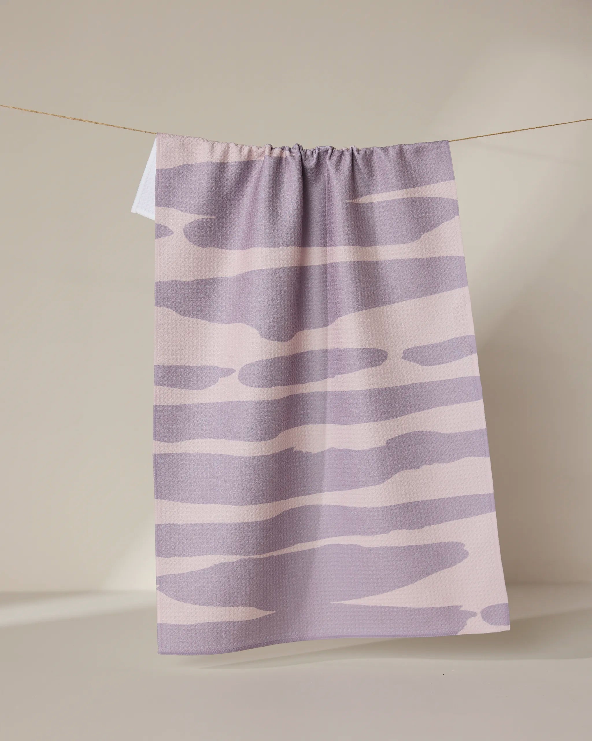The GEOMETRY Reflected - Sunset tea towel, featuring pastel purple and cream abstract wavy stripes and made from recycled materials, hangs on a clothesline against a neutral background.