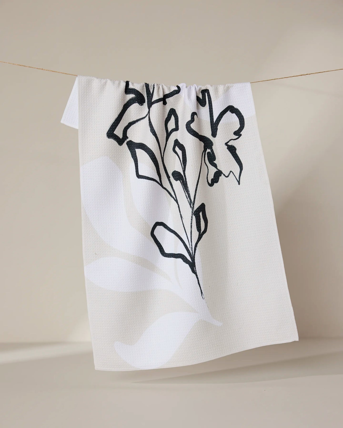 The GEOMETRY Reflected tea towel, made from recycled materials, features a minimalist black and gray botanical design and is shown hanging on a thin string against a neutral background.