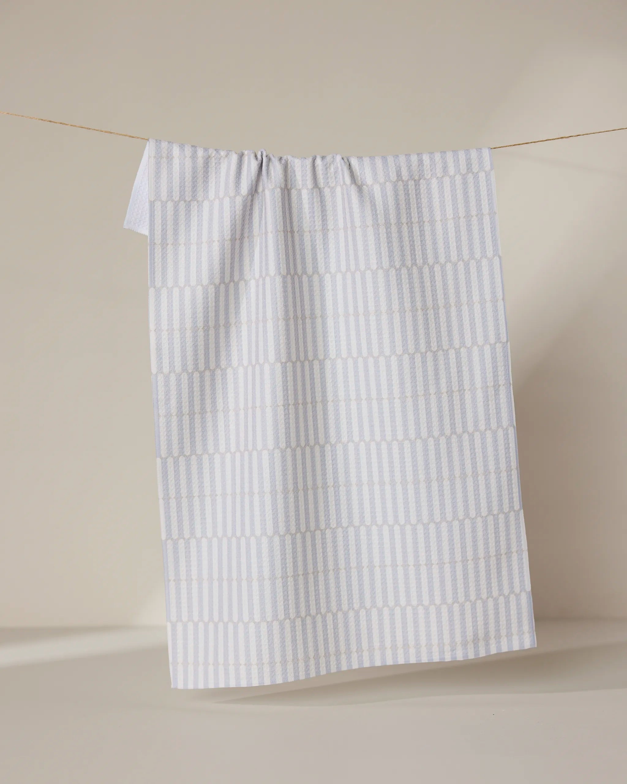 The GEOMETRY Rideaux Neutre tea towel, featuring a subtle light grey geometric pattern on white, is draped over a string against a neutral beige background.