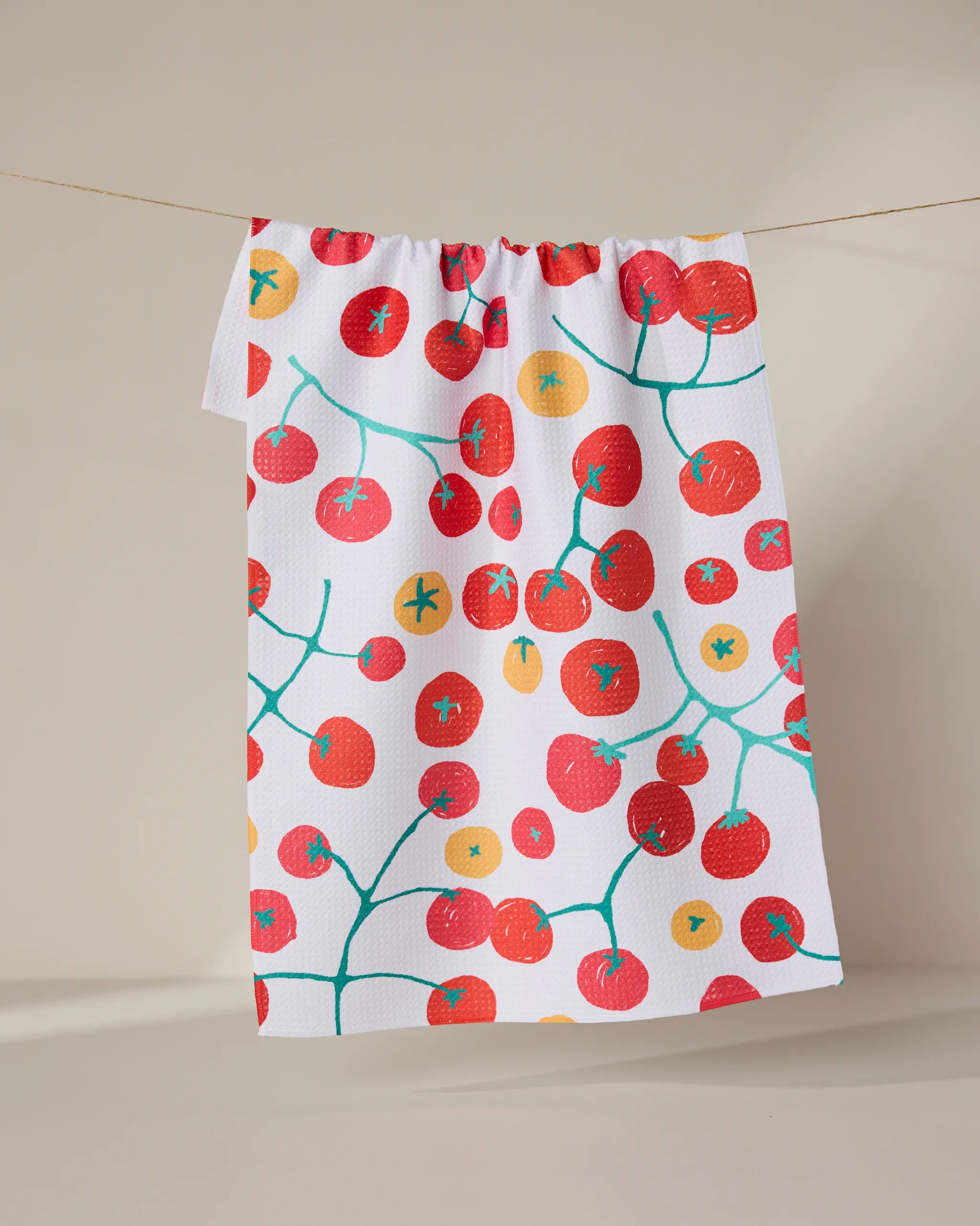 The GEOMETRY Ripe recycled kitchen towel, featuring vibrant red and orange cherry tomatoes on green vines, hangs on a clothesline against a neutral background.