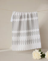 The GEOMETRY Salle Dor kitchen tea towel, featuring a repeating gray feather pattern on white and made from recycled materials, hangs on a string against a beige background with a white flower and green leaves at the bottom right.