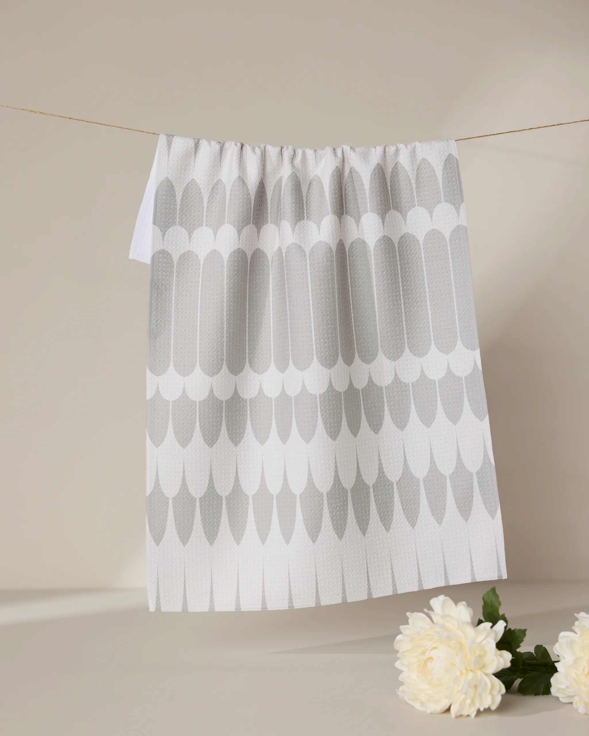 The GEOMETRY Salle Dor kitchen tea towel, featuring a repeating gray feather pattern on white and made from recycled materials, hangs on a string against a beige background with a white flower and green leaves at the bottom right.