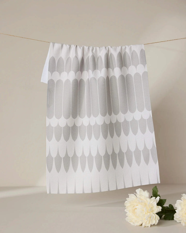 The GEOMETRY Salle Dor kitchen tea towel, featuring a repeating gray feather pattern on white and made from recycled materials, hangs on a string against a beige background with a white flower and green leaves at the bottom right.