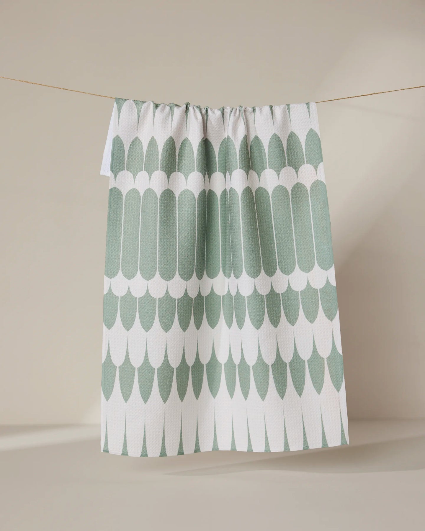 The GEOMETRY Salle Verte tea towel, featuring a green and white scalloped pattern, hangs on a clothesline against a neutral beige background.