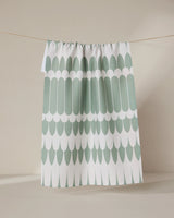 The GEOMETRY Salle Verte tea towel, featuring a green and white scalloped pattern, hangs on a clothesline against a neutral beige background.