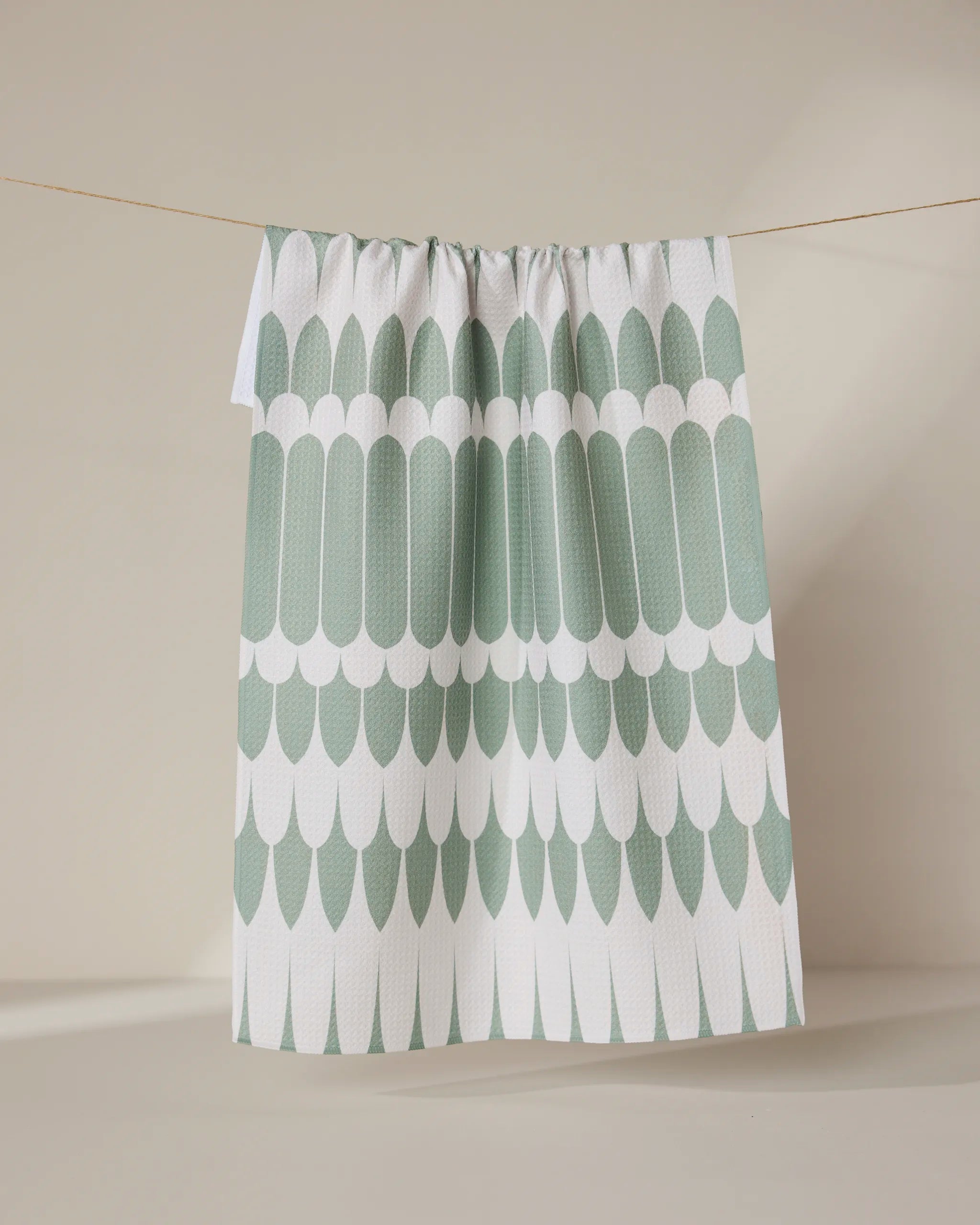 The GEOMETRY Salle Verte tea towel, featuring a green and white scalloped pattern, hangs on a clothesline against a neutral beige background.