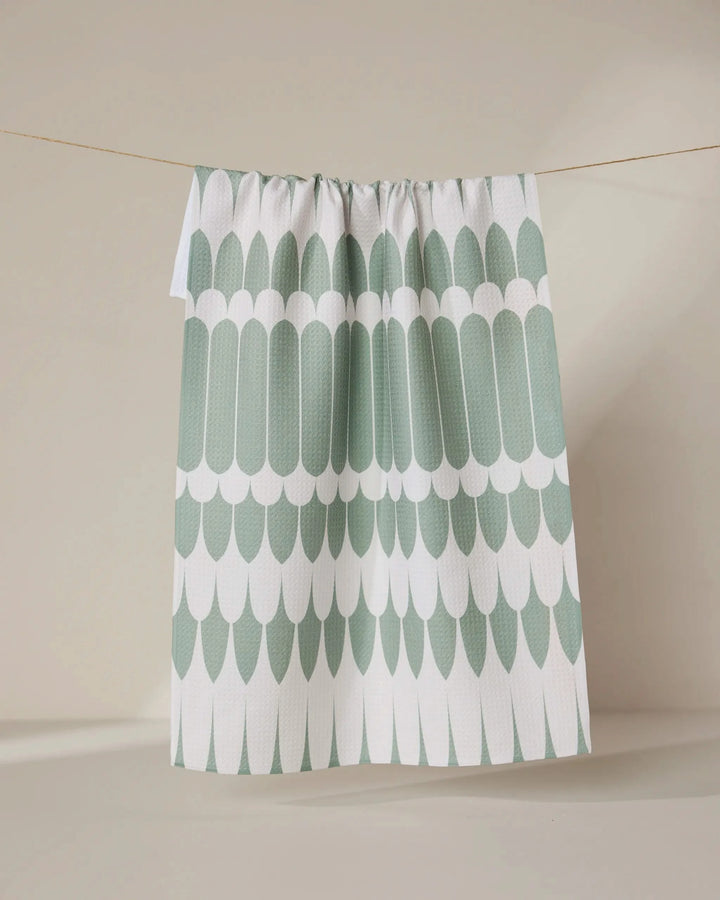 The GEOMETRY Salle Verte tea towel, featuring a green and white scalloped pattern, hangs on a clothesline against a neutral beige background.