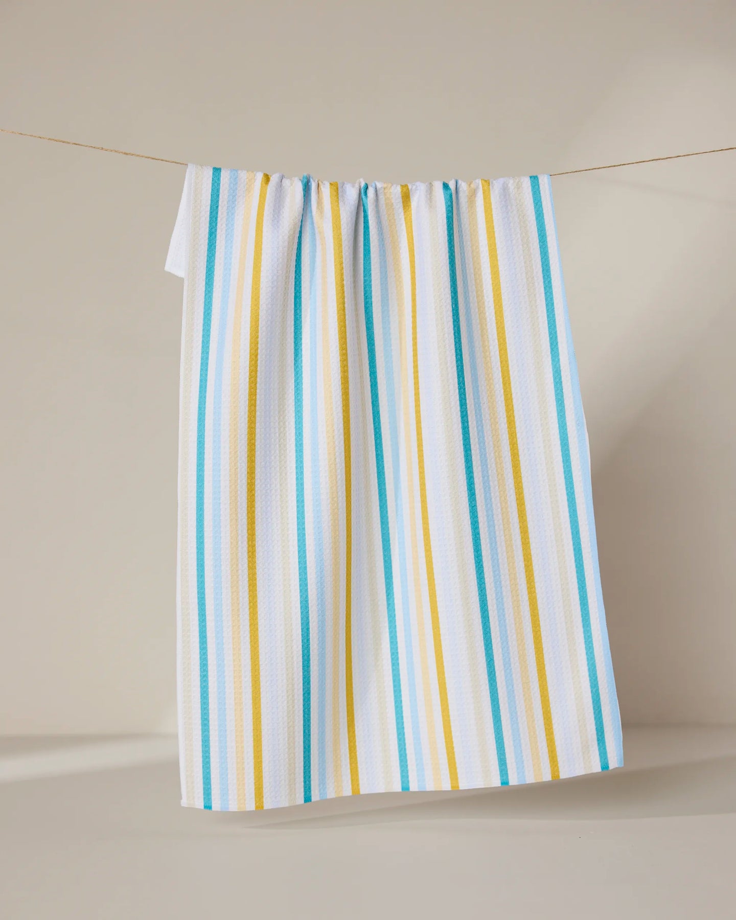 The GEOMETRY Seaside Sunshine tea towel, featuring blue and yellow vertical stripes on white, hangs on a string against a plain, light-colored background.