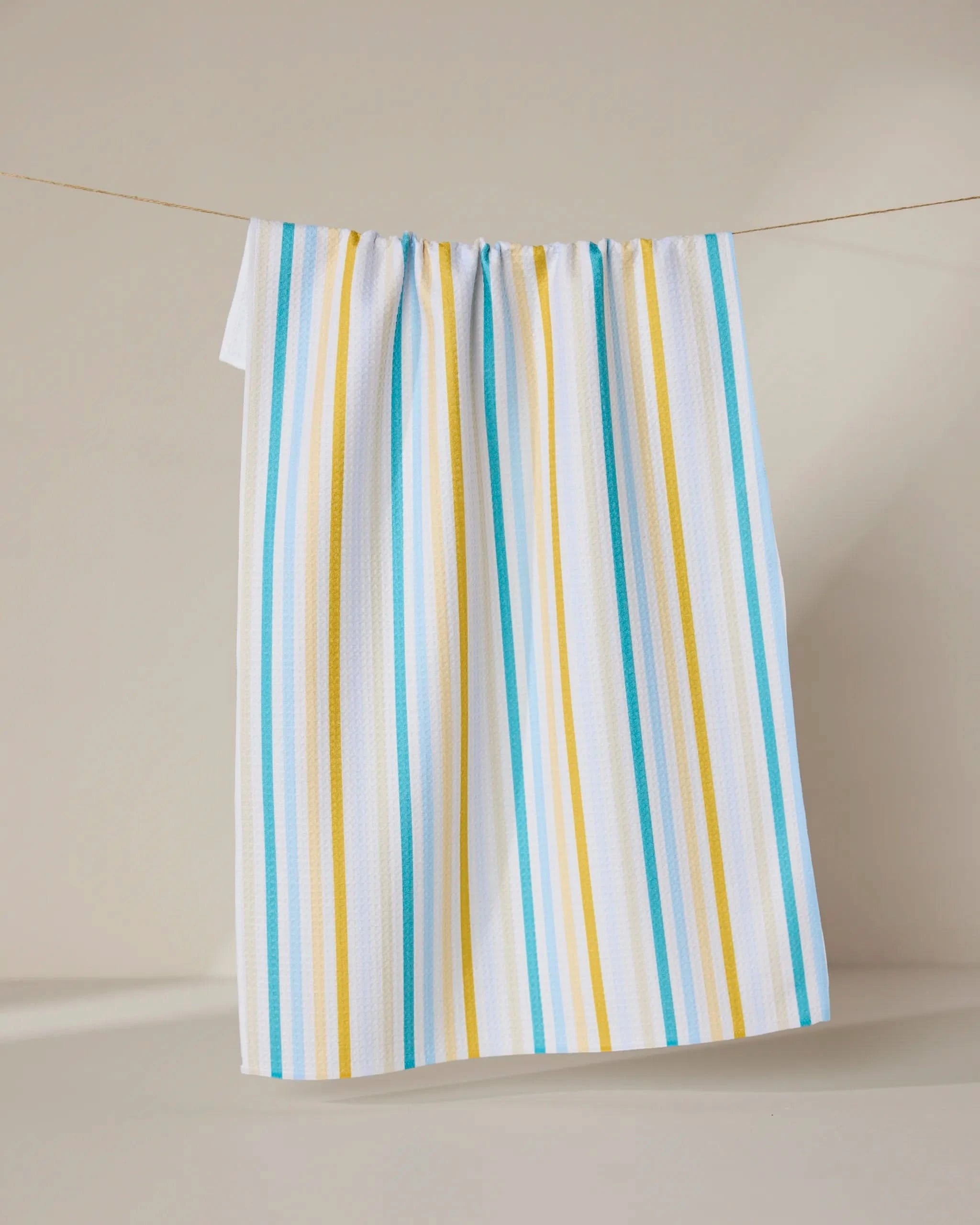 The GEOMETRY Seaside Sunshine tea towel, featuring blue and yellow vertical stripes on white, hangs on a string against a plain, light-colored background.