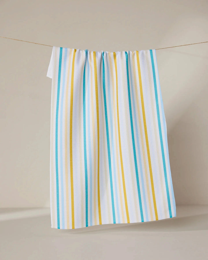 The GEOMETRY Seaside Sunshine tea towel, featuring blue and yellow vertical stripes on white, hangs on a string against a plain, light-colored background.