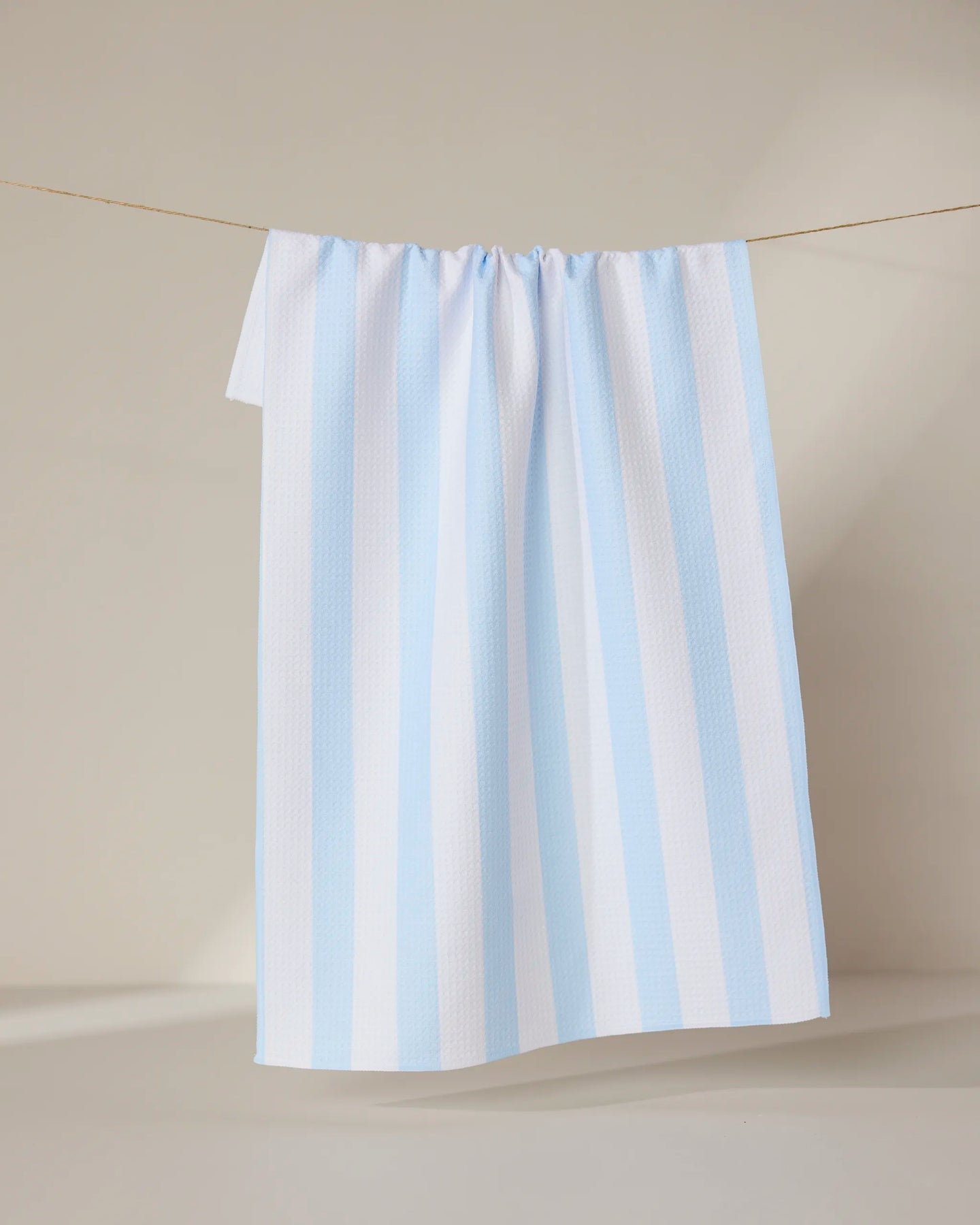 The GEOMETRY Seaside Stripes towel, featuring ultra-absorbent fabric and vertical light blue and white stripes, hangs neatly on a clothesline against a neutral background.