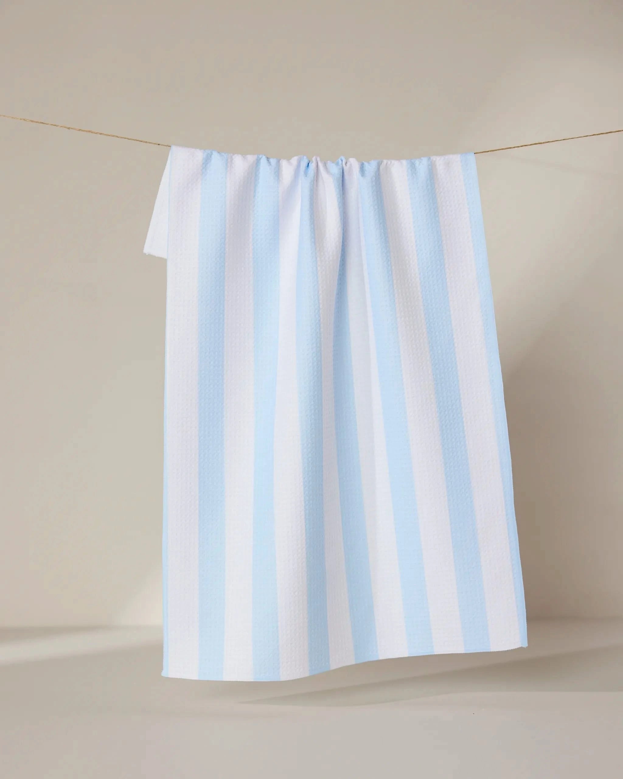 The GEOMETRY Seaside Stripes towel, featuring ultra-absorbent fabric and vertical light blue and white stripes, hangs neatly on a clothesline against a neutral background.