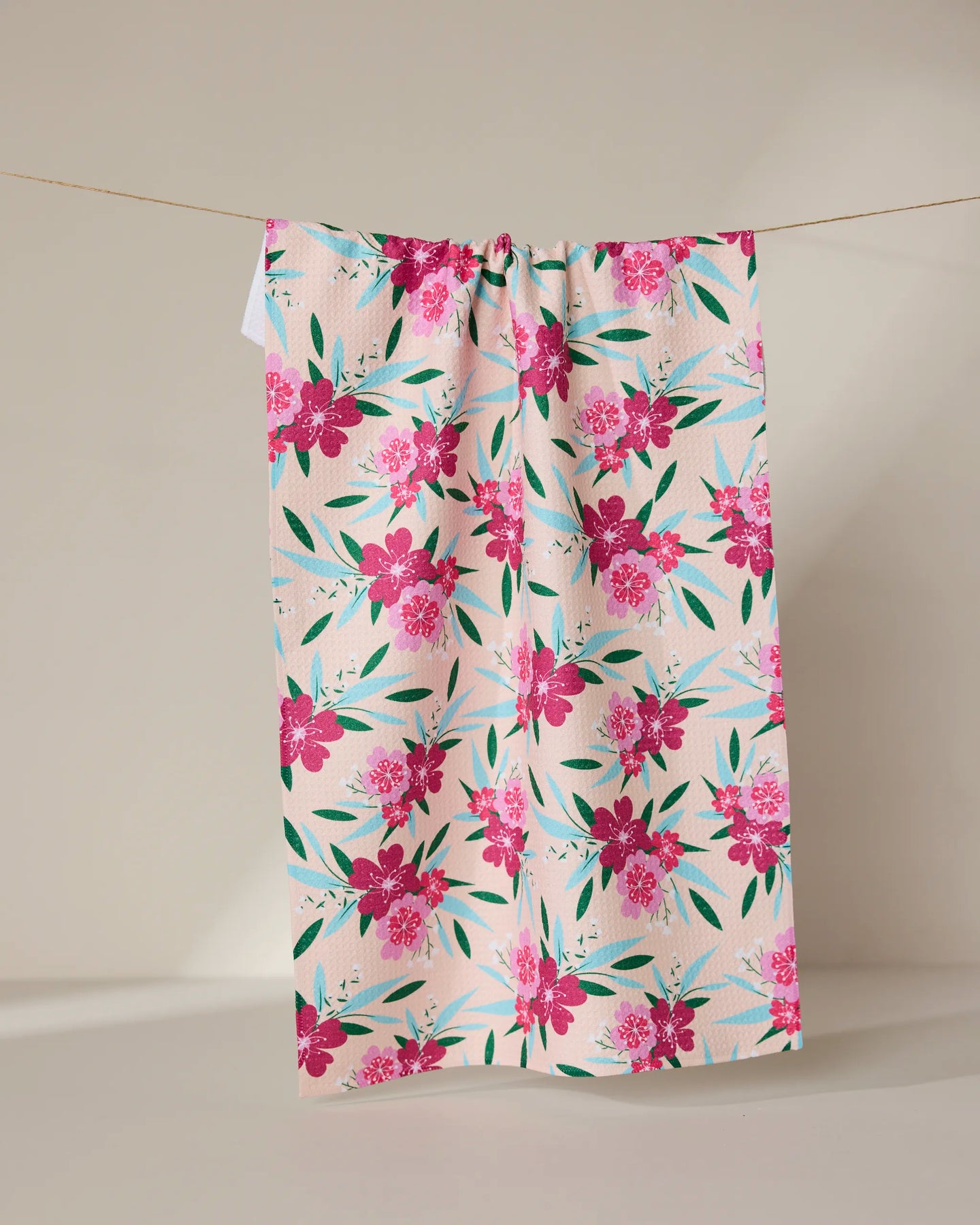 The GEOMETRY Sending Love tea towel, crafted from recycled materials and featuring pink flowers with green leaves, hangs on a clothesline against a plain backdrop.