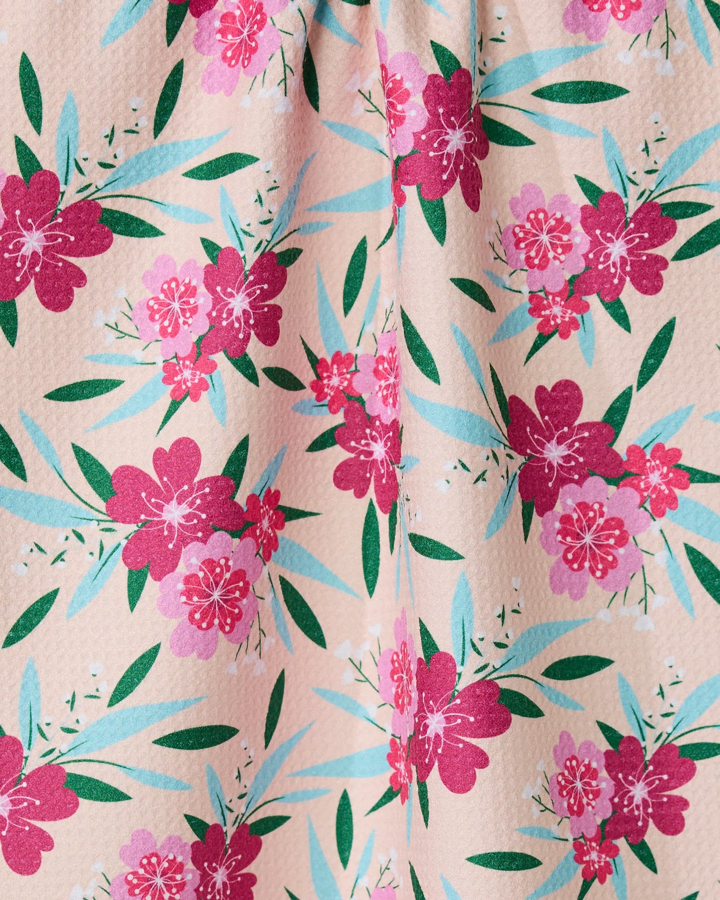 A close-up of GEOMETRYs Sending Love tea towel, made from recycled materials, features red and pink flowers, green leaves, and blue-green foliage on a pink background. The textured fabric is slightly gathered at the top.