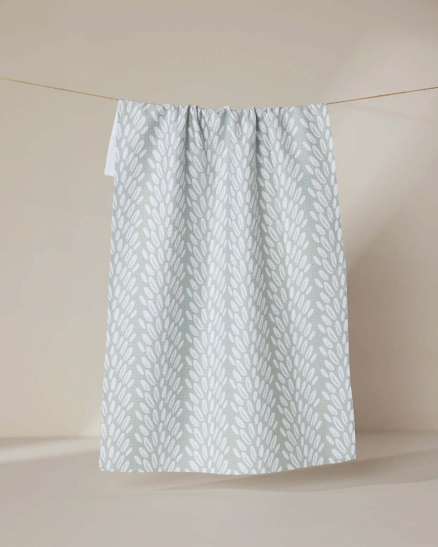 The GEOMETRY Seed tea towel, featuring a white leafy pattern on light green, hangs on a thin string against a plain beige background.