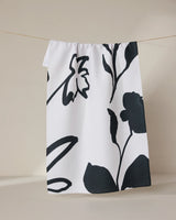The Shade tea towel by GEOMETRY, featuring large bold black floral prints and made from recycled materials, hangs from a thin string against a neutral background.