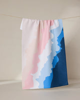 The Shore Tea Towel by GEOMETRY, featuring wavy pastel stripes in pink, white, light blue, and dark blue, hangs on a clothesline. Made with Geoweave fabric, it offers superior absorbency and durability against a neutral background.