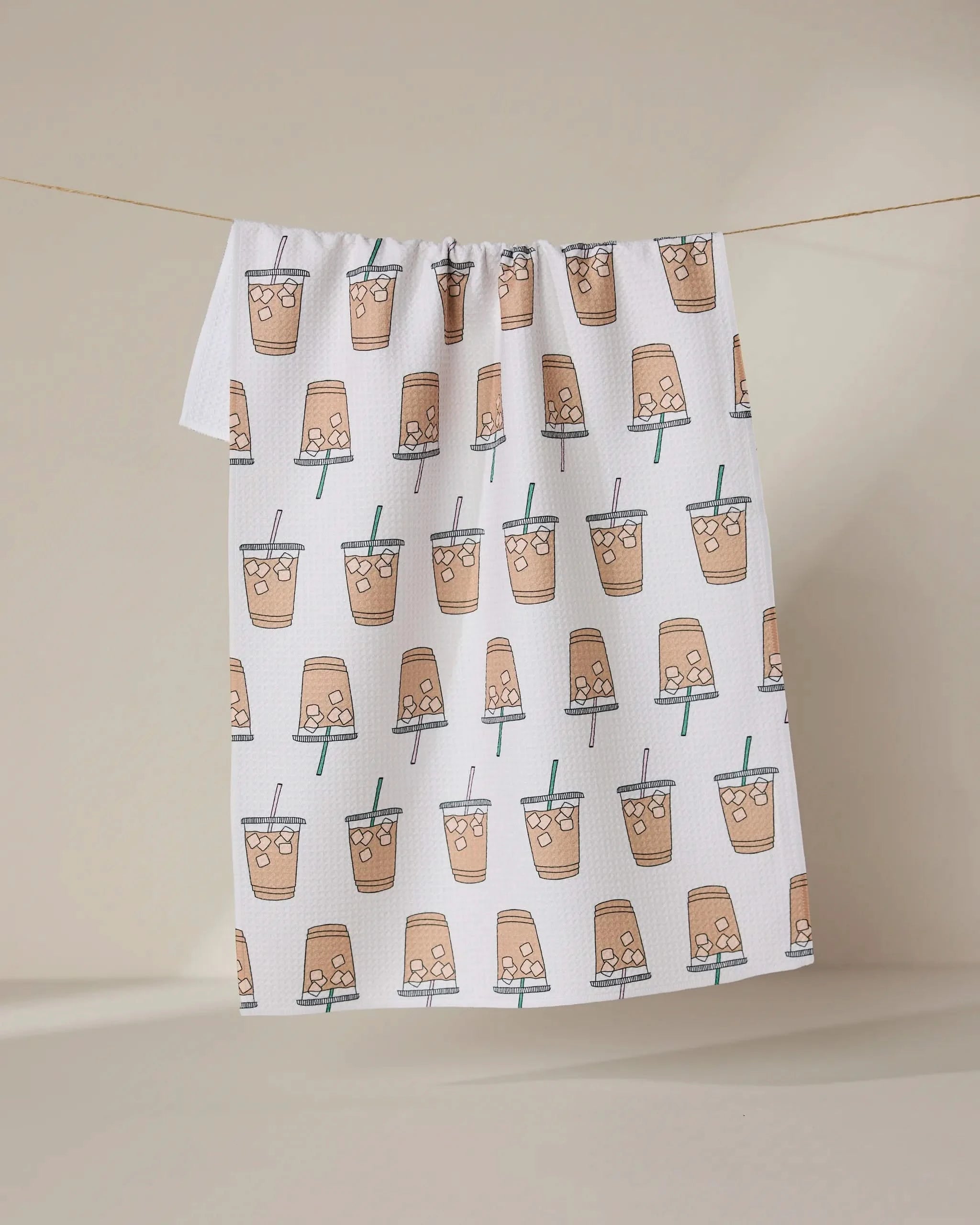 The GEOMETRY Sippin on Summer towel, ultra-absorbent and white with a repeating illustrated bubble tea cup pattern, hangs on a clothesline against a neutral background.