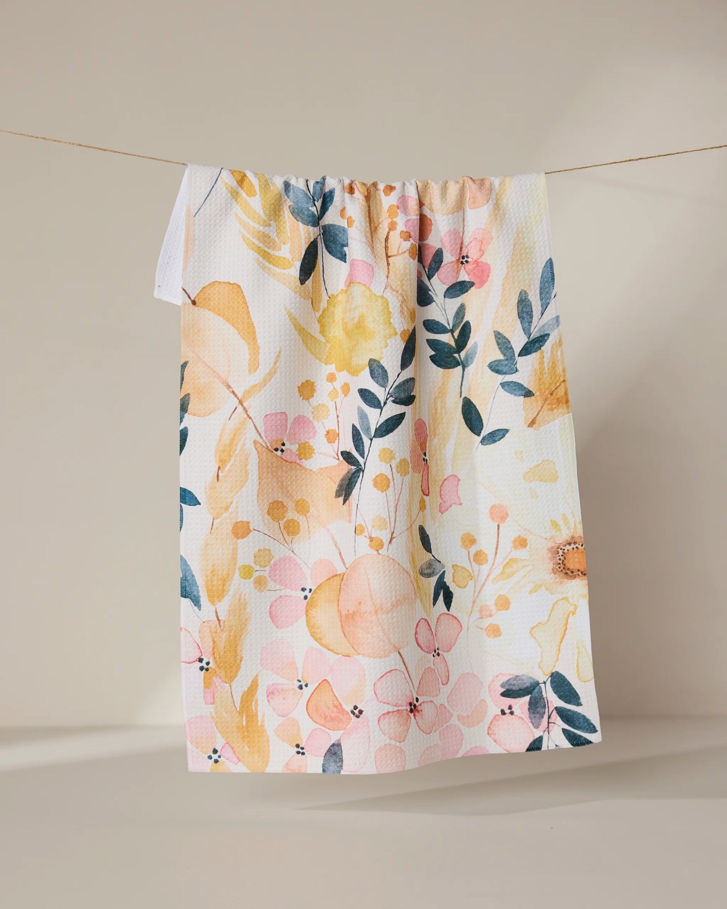 The GEOMETRY Sleepy Fields white kitchen towel, featuring a vibrant floral pattern in yellow, pink, and blue and made from recycled materials, hangs on a string against a neutral beige background.