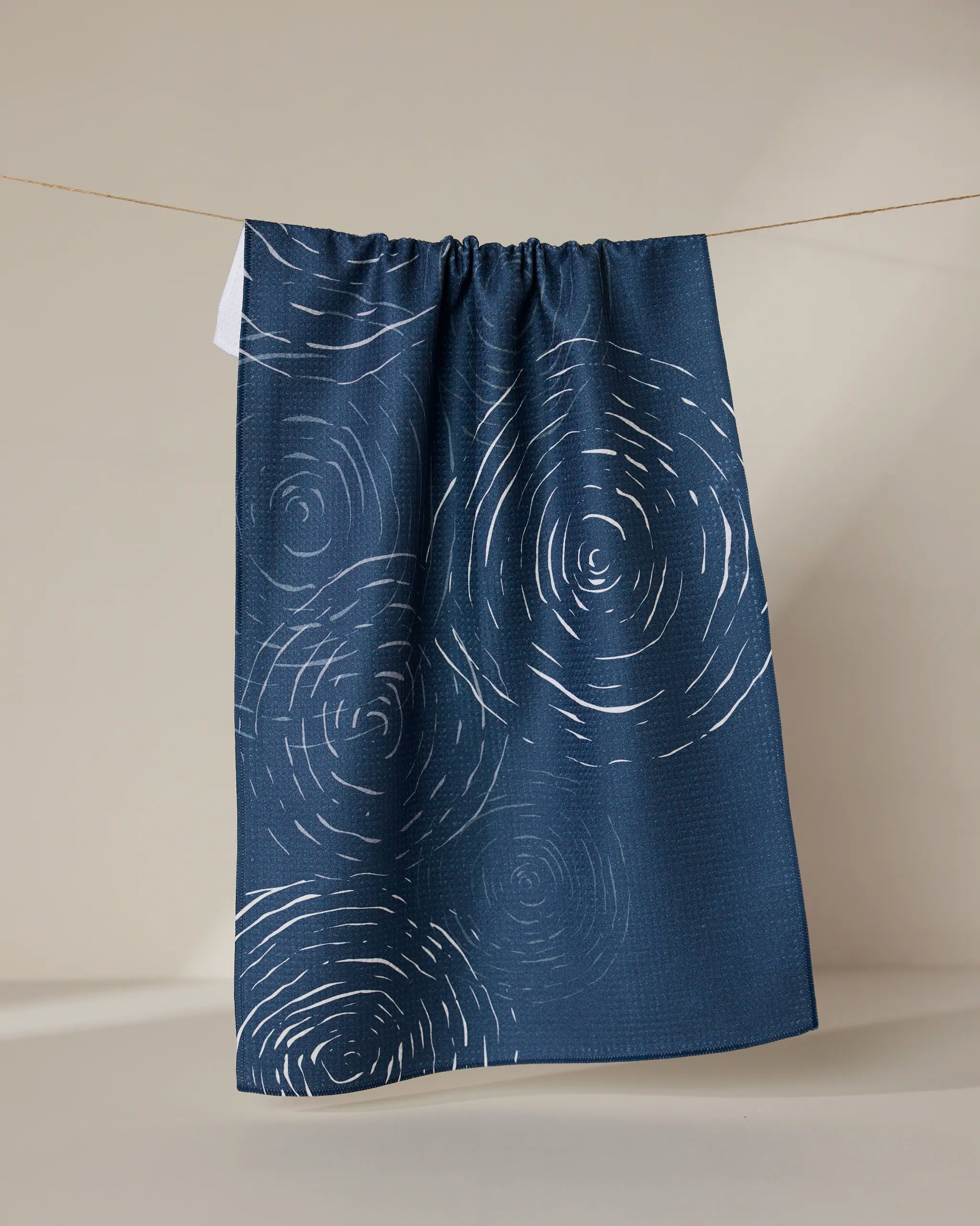The GEOMETRY Soft Steps - Midnight kitchen towel, made from recycled materials and featuring swirling white lines on navy blue, hangs on a string against a beige background.