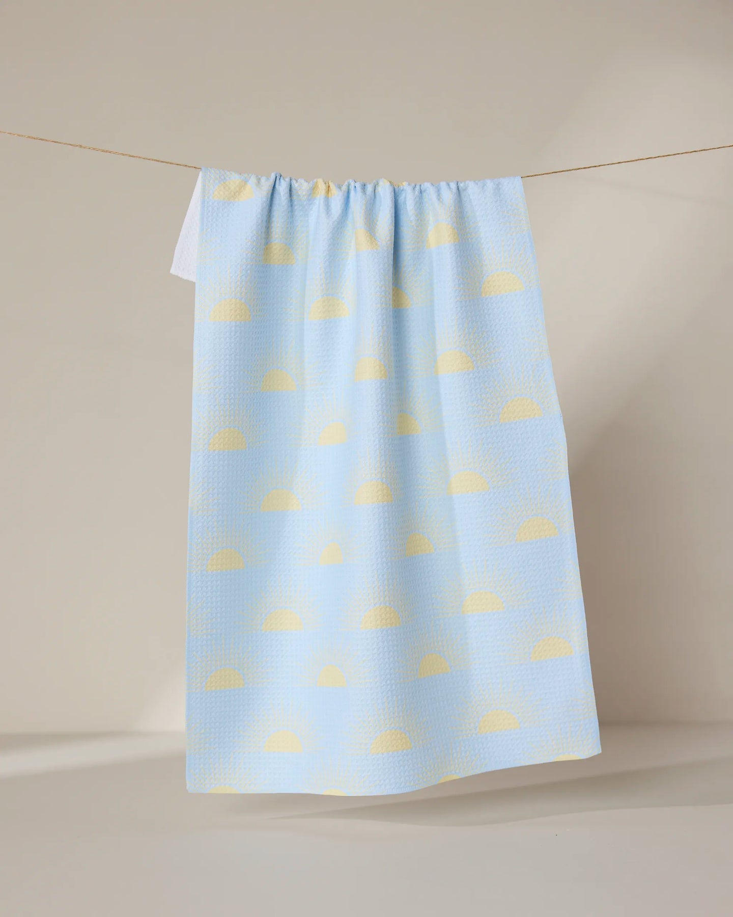 The Soft Summer Sun tea towel by GEOMETRY, made from recycled materials and decorated with yellow semicircles on pastel blue, hangs from a clothesline against a neutral background.