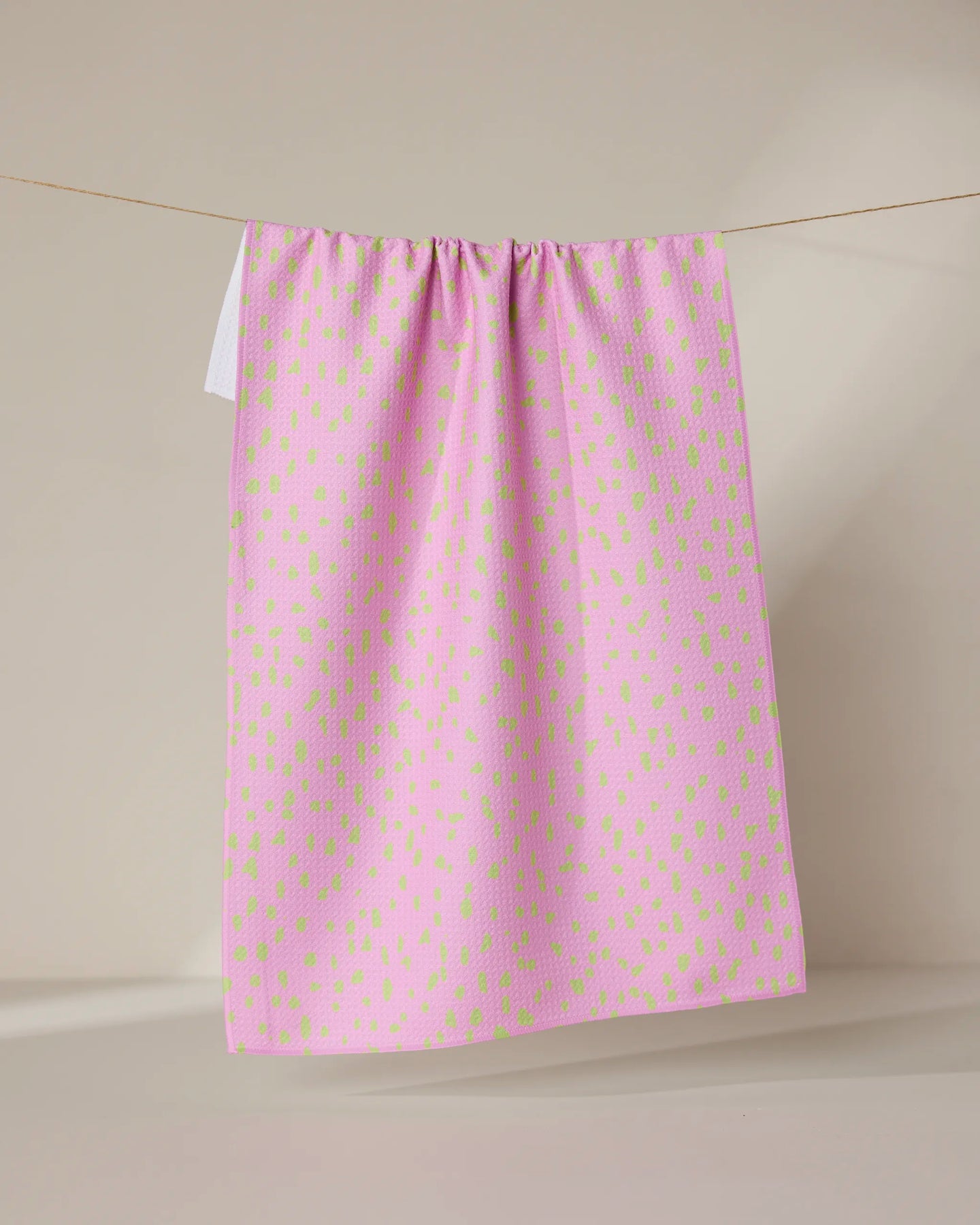 The GEOMETRY Speckle Pink tea towel, featuring ultra-absorbent fabric and small irregular green spots, hangs draped over a clothesline against a plain beige background.