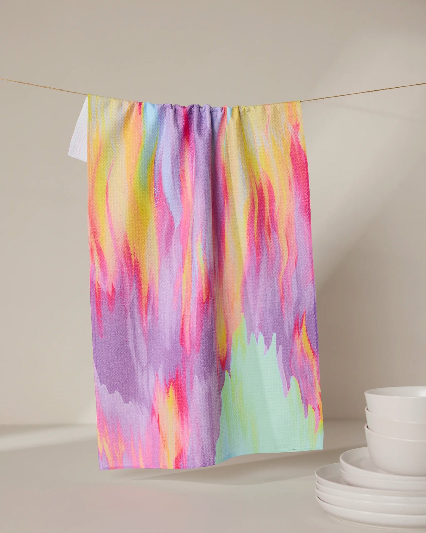 The Splash of Summer tea towel by GEOMETRY, featuring a quick-drying abstract pattern in pink, yellow, purple, and light green, hangs above stacked white bowls on a neutral background.