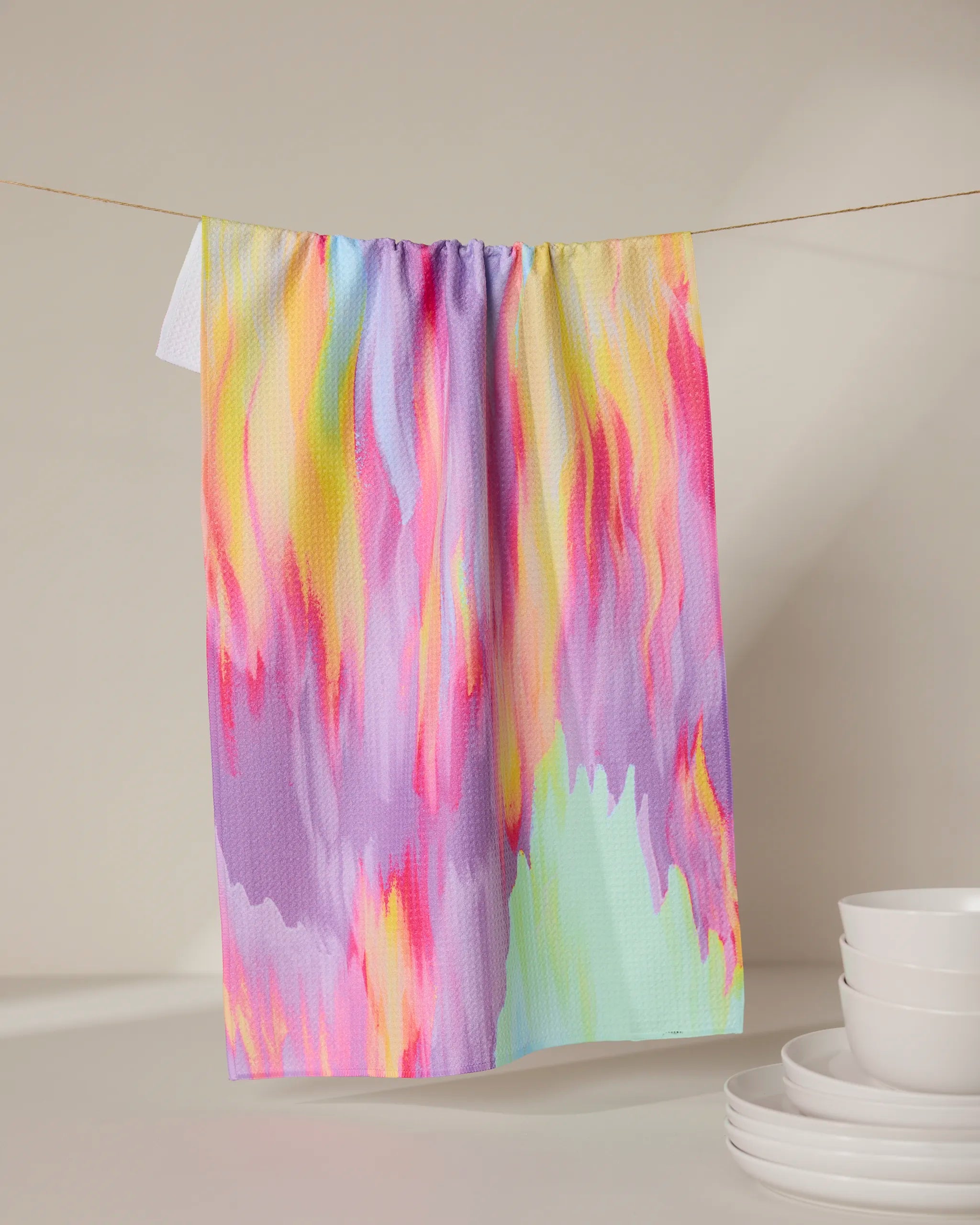 The Splash of Summer tea towel by GEOMETRY, featuring a quick-drying abstract pattern in pink, yellow, purple, and light green, hangs above stacked white bowls on a neutral background.