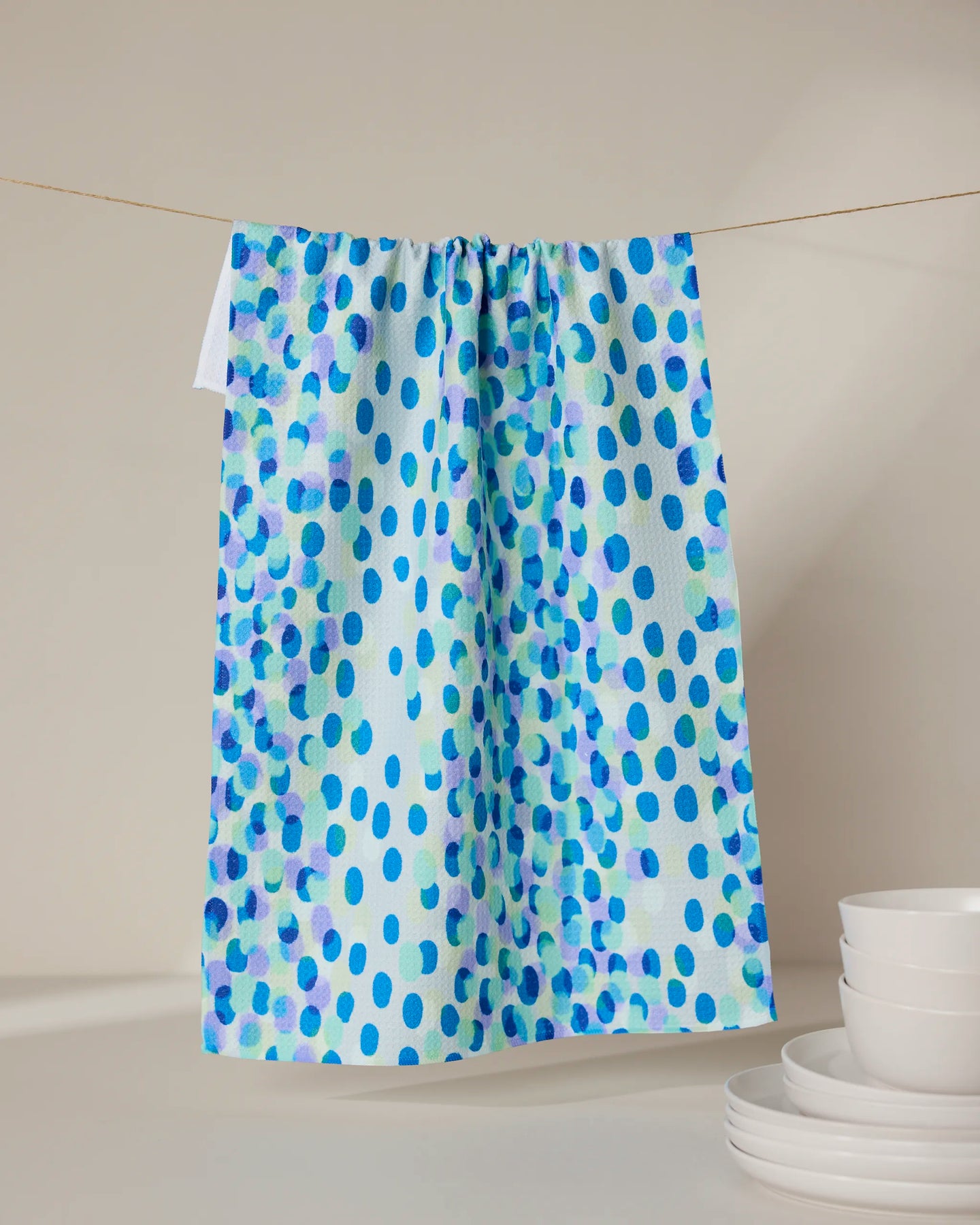 The GEOMETRY Spotty Summer kitchen towel, featuring blue and purple polka dots, hangs on a string against a neutral background with a stack of white plates to the right.
