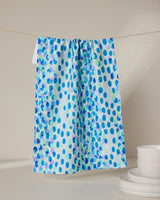 The GEOMETRY Spotty Summer kitchen towel, featuring blue and purple polka dots, hangs on a string against a neutral background with a stack of white plates to the right.
