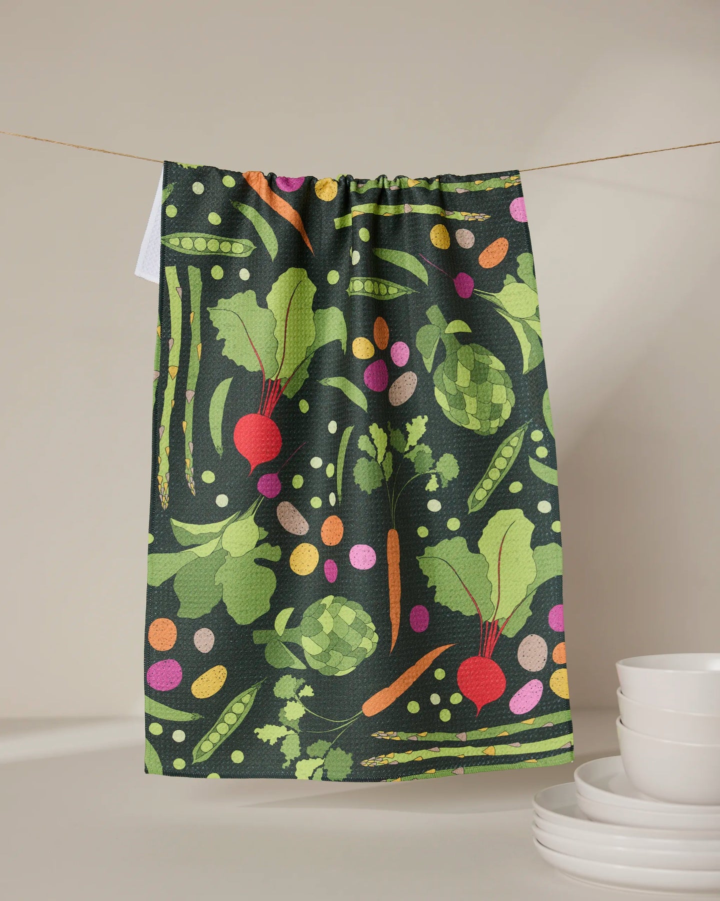 The GEOMETRY Spring Sprout tea towel, featuring a vibrant veggie pattern—beets, peas, artichokes, carrots, and leafy greens—crafted from recycled materials, hangs above stacked white bowls and plates against a neutral backdrop.