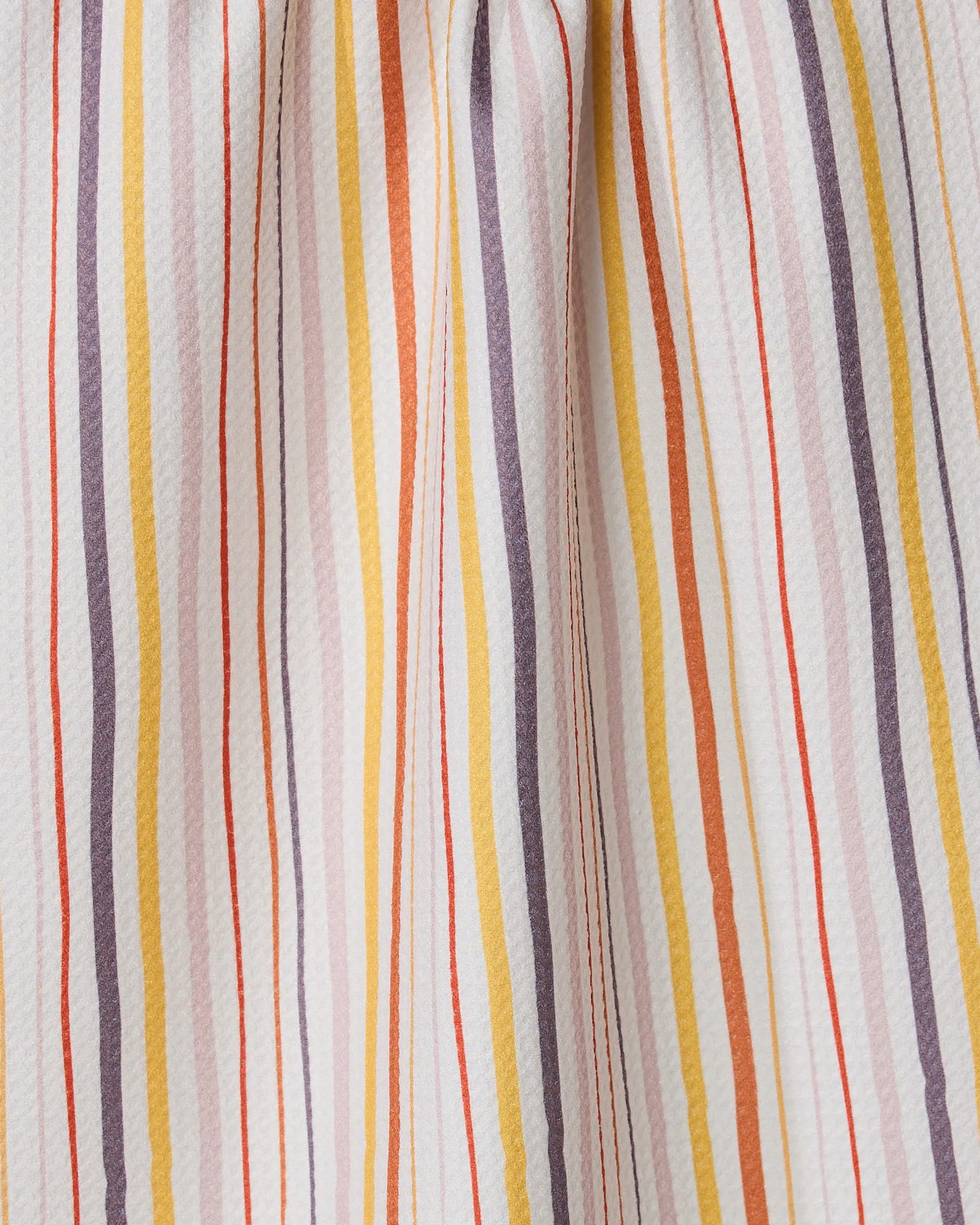 A close-up of GEOMETRY’s Stripe Season towel shows ultra-absorbent fabric with vertical yellow, orange, lavender, and red stripes on white. The towel is gently gathered, creating soft folds and shadows.