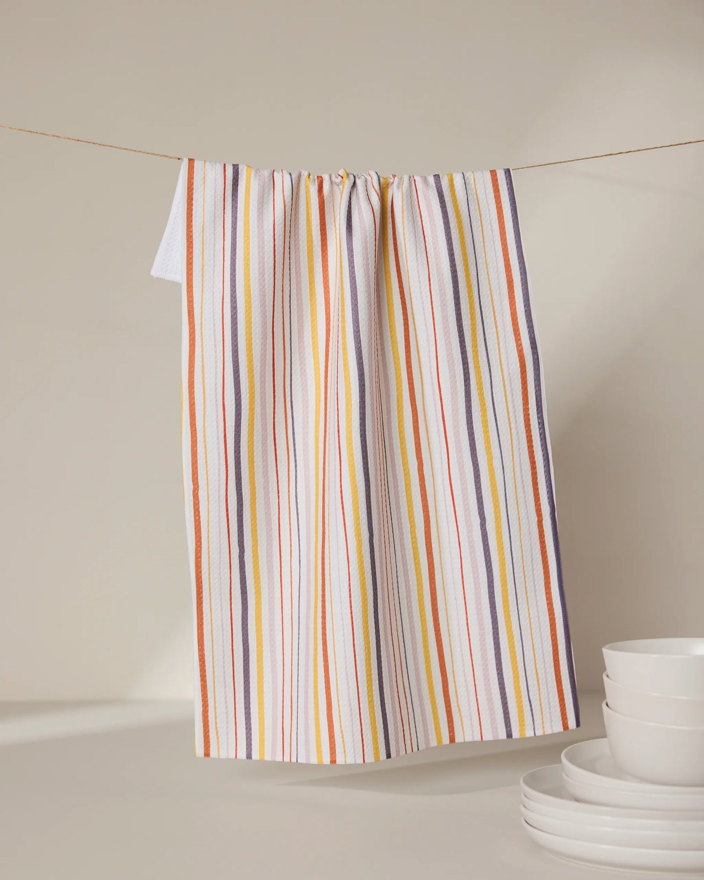 The GEOMETRY Stripe Season tea towel, featuring orange, yellow, purple, and white stripes, hangs on a clothesline behind stacked white bowls and plates. Ultra-absorbent, it adds vibrant color and function to any neutral kitchen.