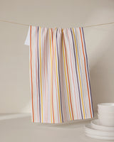 The GEOMETRY Stripe Season tea towel, featuring orange, yellow, purple, and white stripes, hangs on a clothesline behind stacked white bowls and plates. Ultra-absorbent, it adds vibrant color and function to any neutral kitchen.