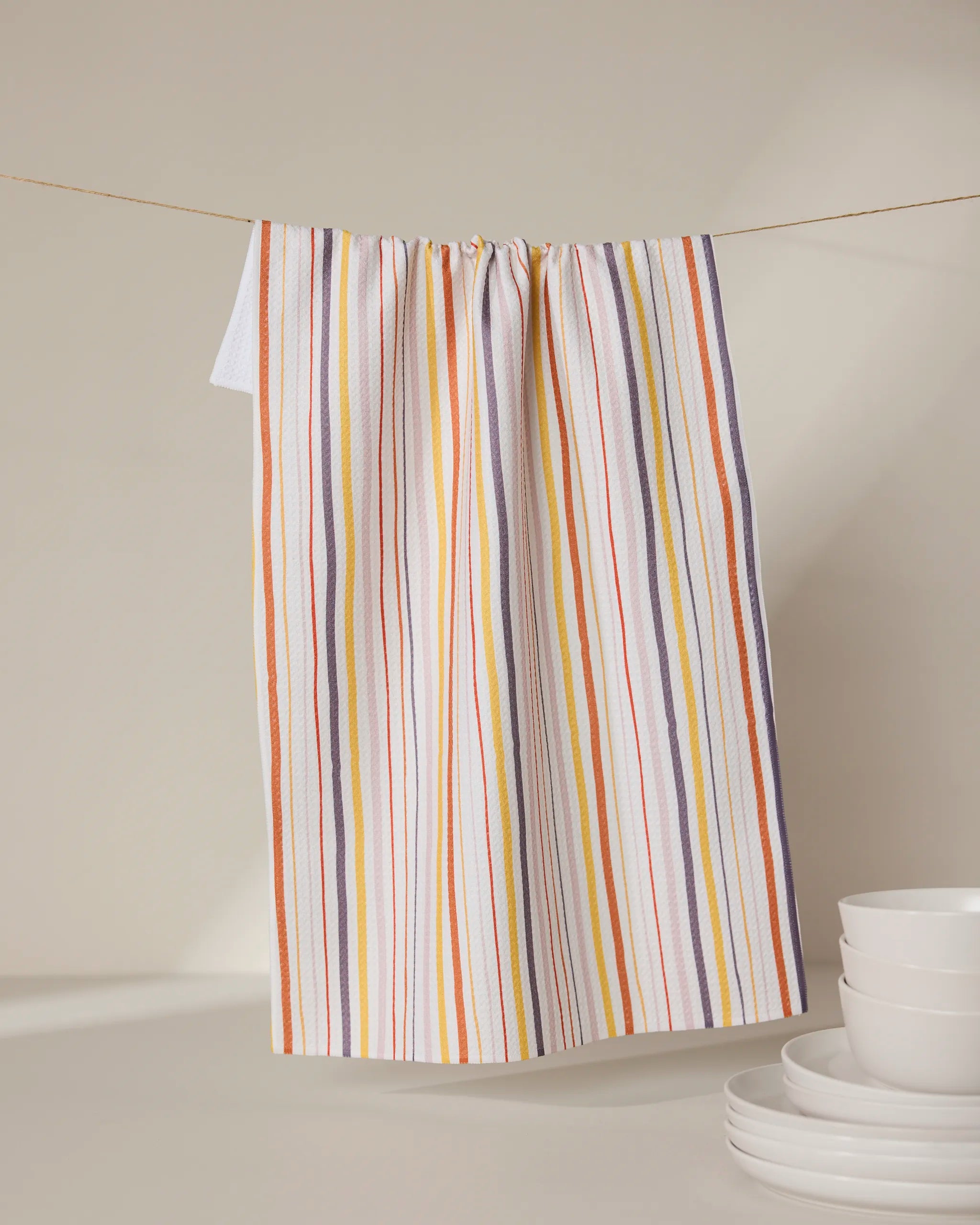The GEOMETRY Stripe Season tea towel, featuring orange, yellow, purple, and white stripes, hangs on a clothesline behind stacked white bowls and plates. Ultra-absorbent, it adds vibrant color and function to any neutral kitchen.
