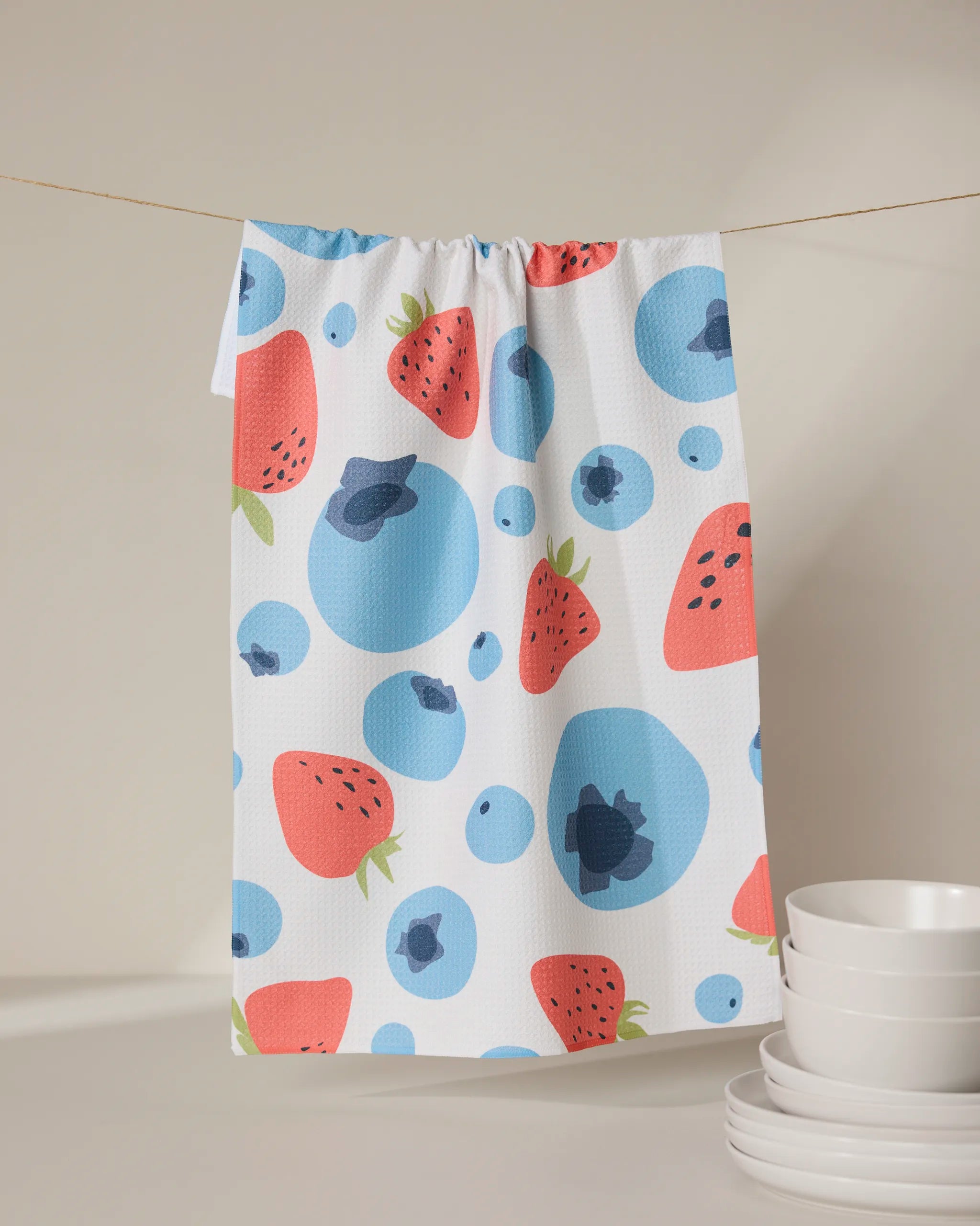 A GEOMETRY Sunny Berries recycled kitchen towel, featuring a vibrant strawberry and blueberry pattern, hangs on a string above stacked white bowls.