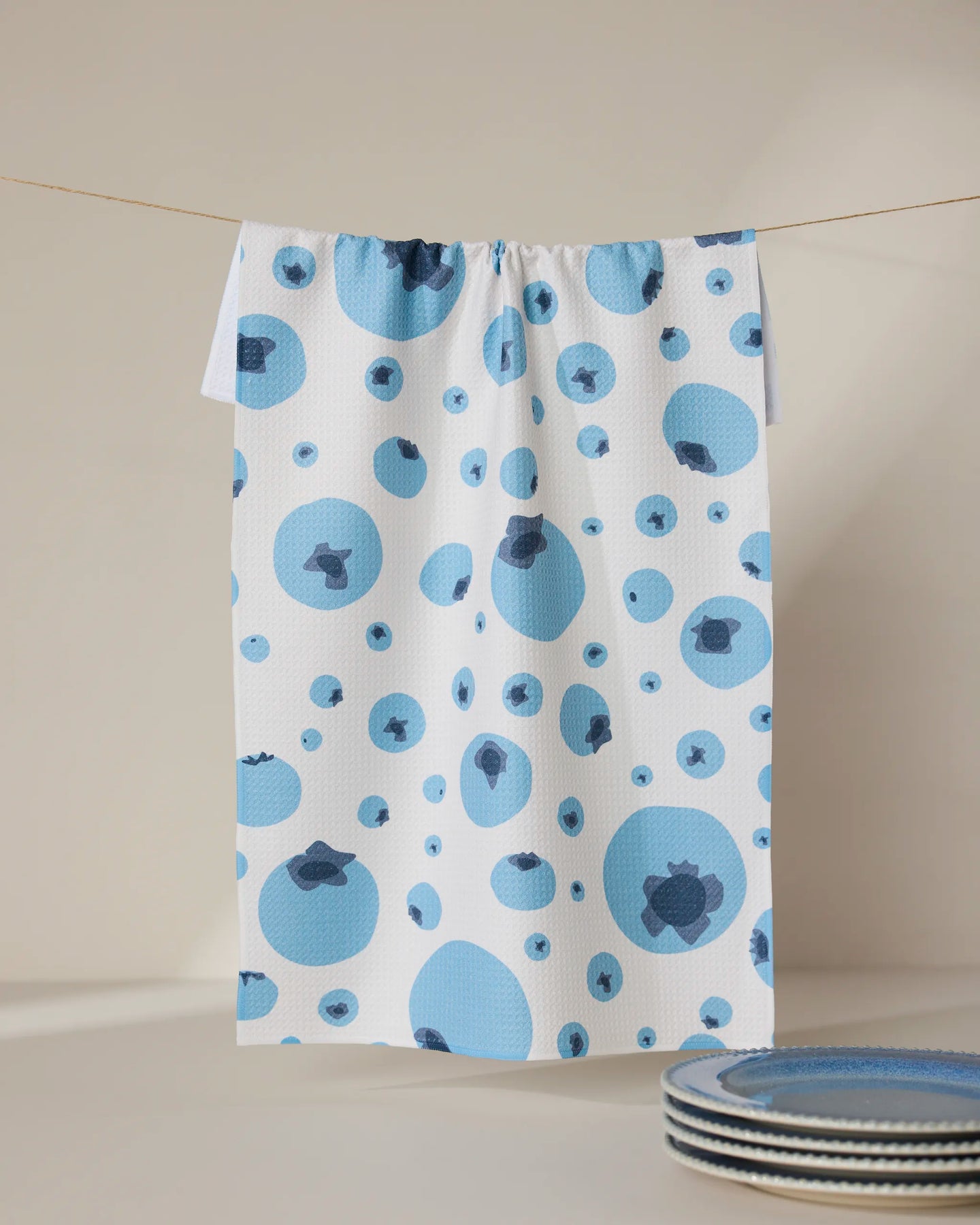 The GEOMETRY Sunny Blueberries white tea towel, crafted from absorbent Geoweave fabric and featuring a blue and dark blue blueberry pattern, hangs on a thin line. Blue-rimmed plates are partially seen in the lower right against a neutral background.