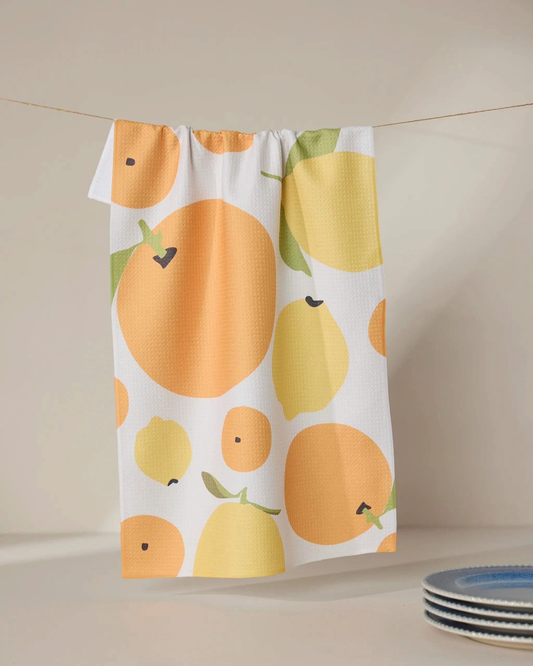 The GEOMETRY “Sunny Lemons And Oranges” white tea towel, featuring a vibrant orange, lemon, and leaf pattern, hangs on a clothesline near a stack of blue and white plates against a softly lit neutral background.
