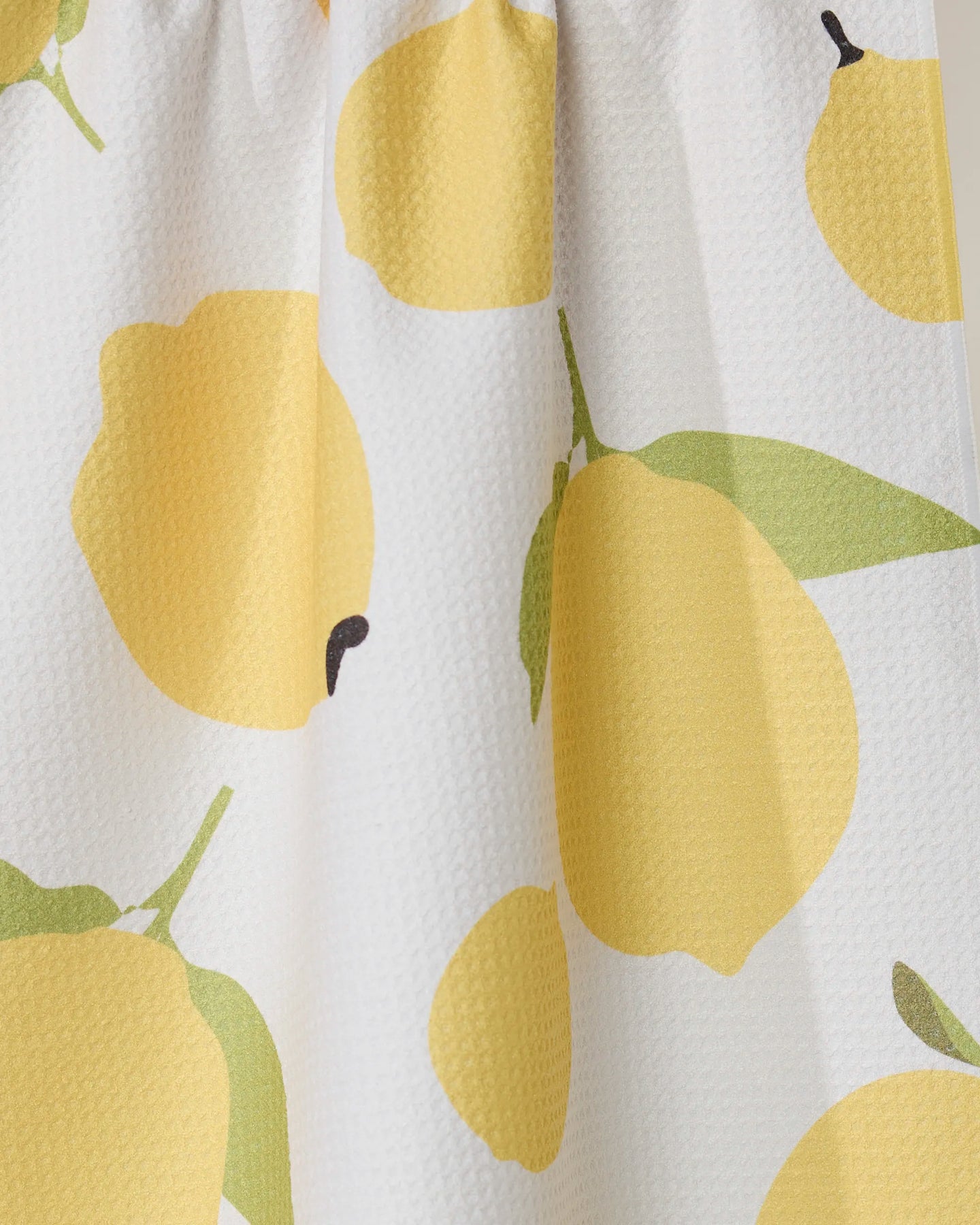 A close-up of the GEOMETRY Sunny Lemons tea towel, made from recycled materials, with white fabric, bold yellow lemons, green leaves, and a gathered seam at the top.