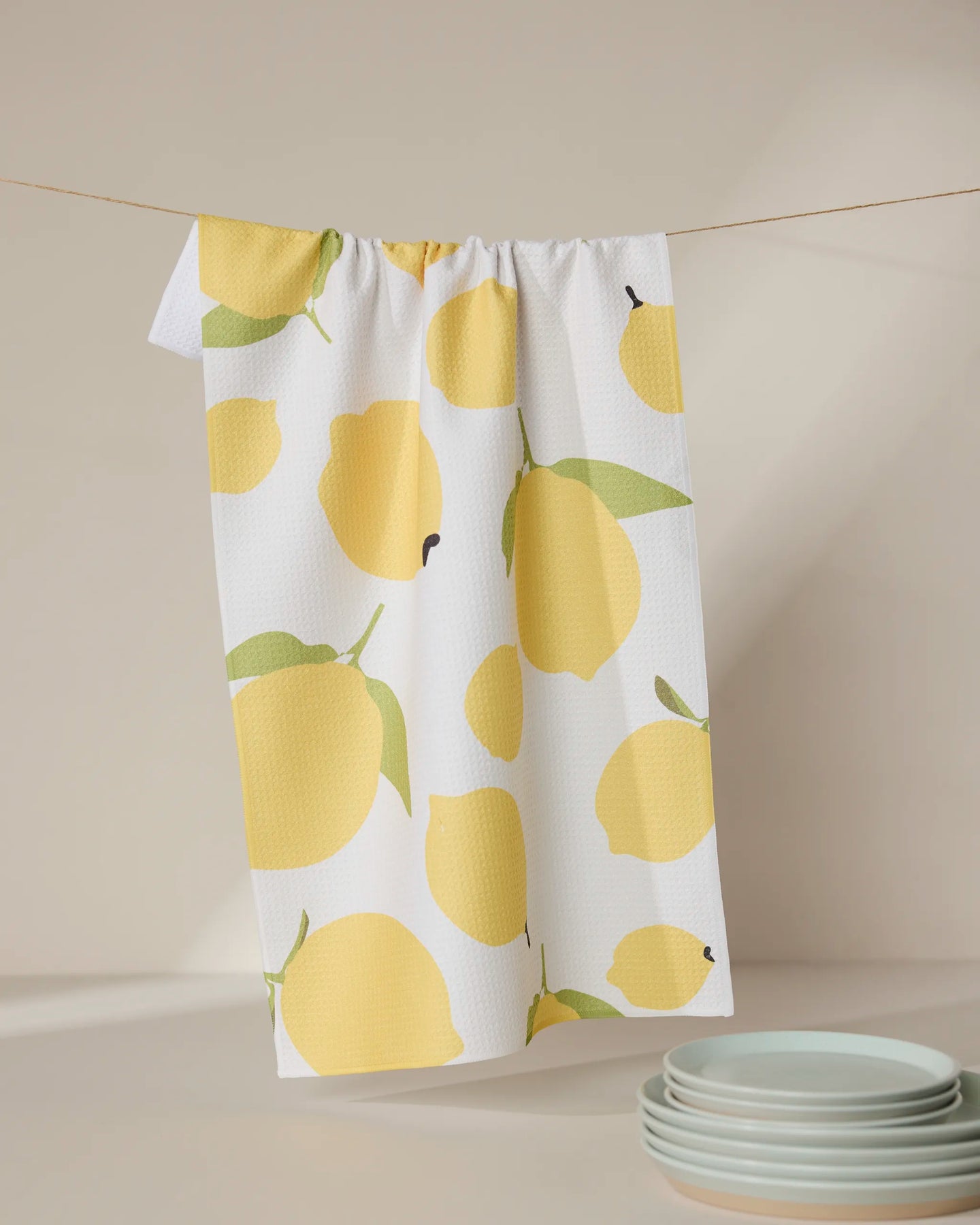The GEOMETRY Sunny Lemons tea towel, crafted from recycled materials and featuring a yellow lemon print, hangs on a line. Light blue plates are stacked on a softly lit, neutral countertop in the corner.