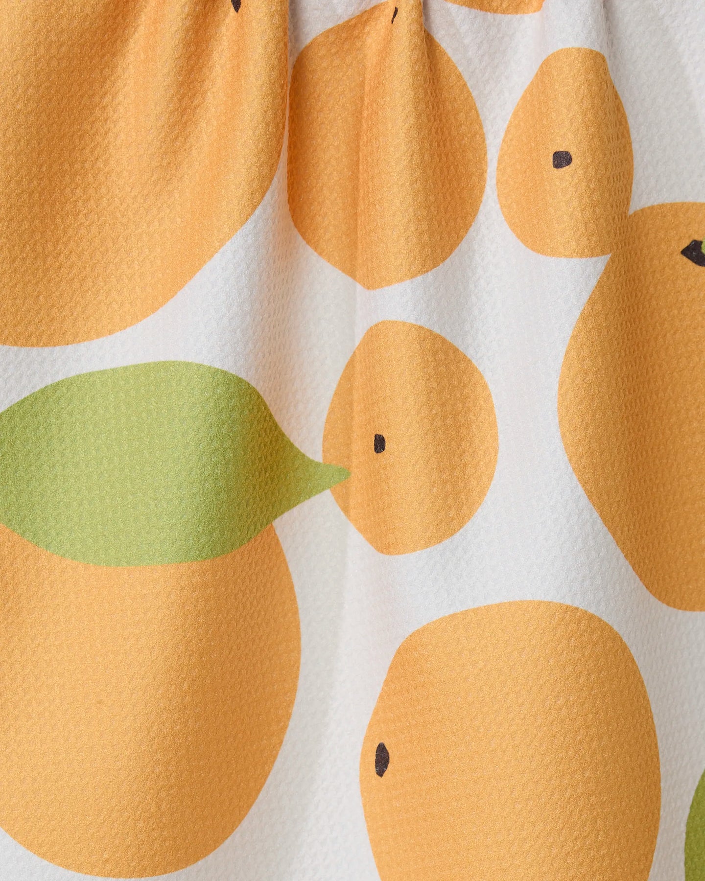 A close-up of GEOMETRYs Sunny Oranges kitchen tea towel, featuring simple orange circles and green leaves on white. Made from recycled materials, the fabric has a slightly textured surface.