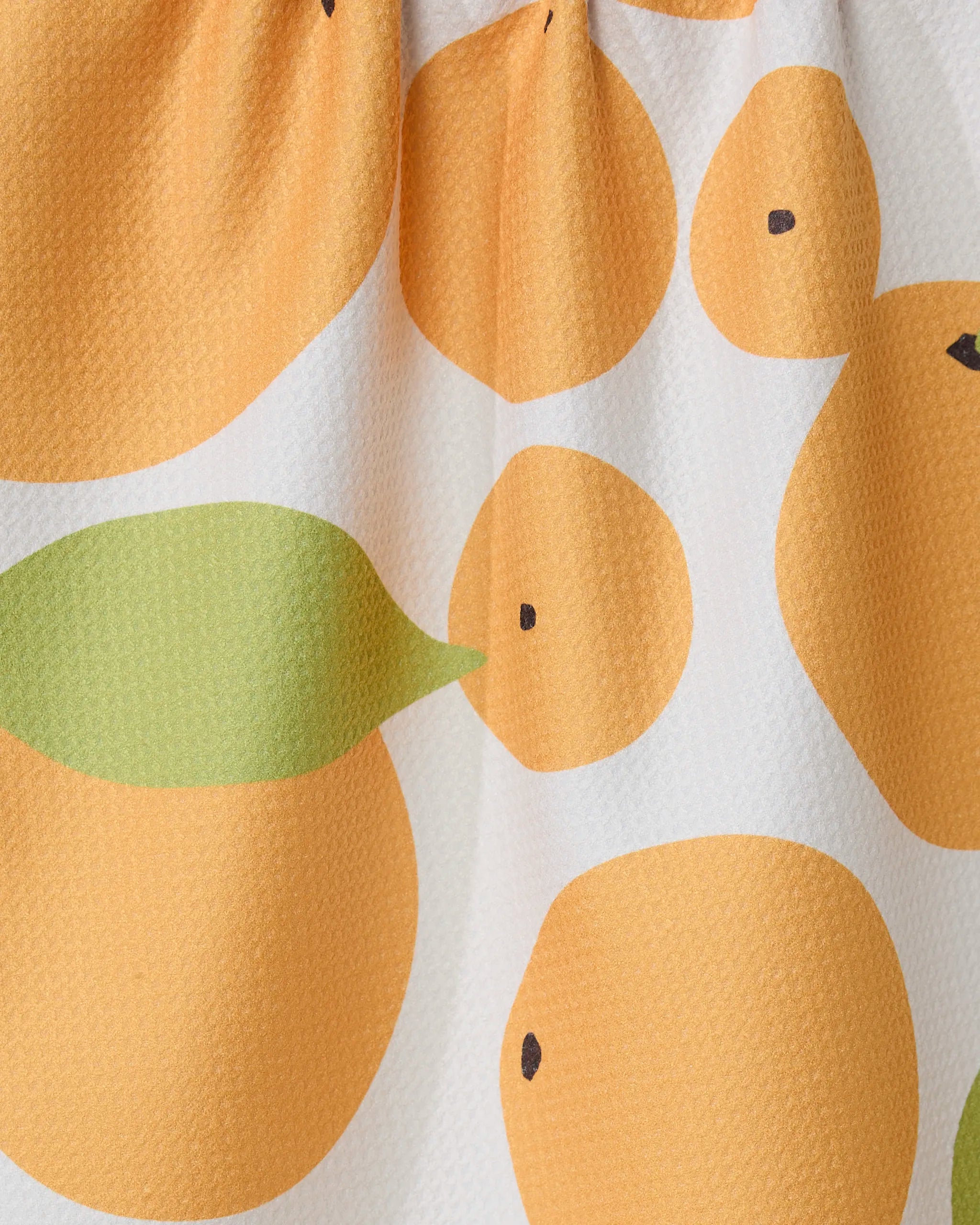A close-up of GEOMETRYs Sunny Oranges kitchen tea towel, featuring simple orange circles and green leaves on white. Made from recycled materials, the fabric has a slightly textured surface.
