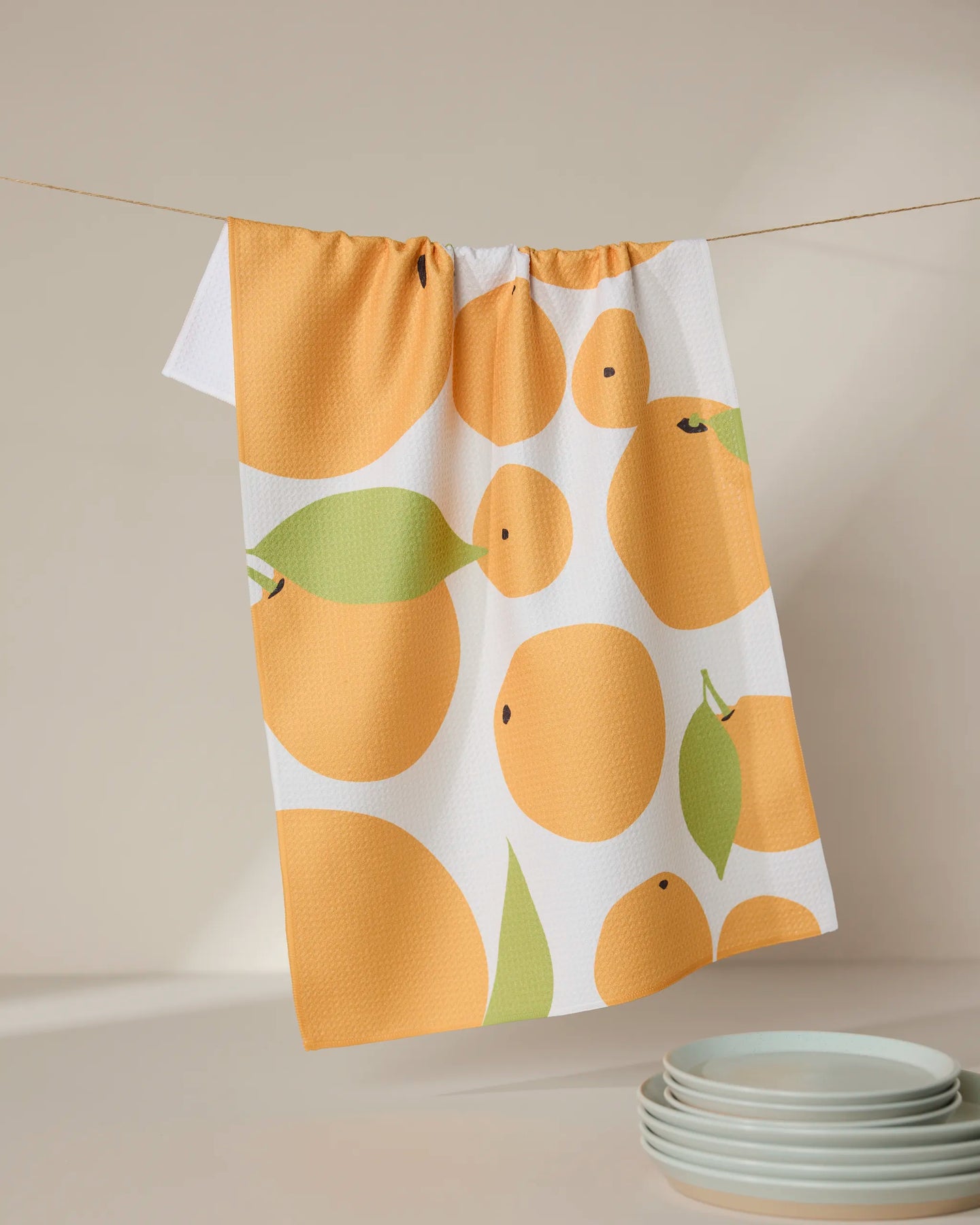 The GEOMETRY Sunny Oranges white kitchen tea towel, crafted from recycled materials, features a bold orange and green fruit print and hangs above stacked white plates on a neutral surface.
