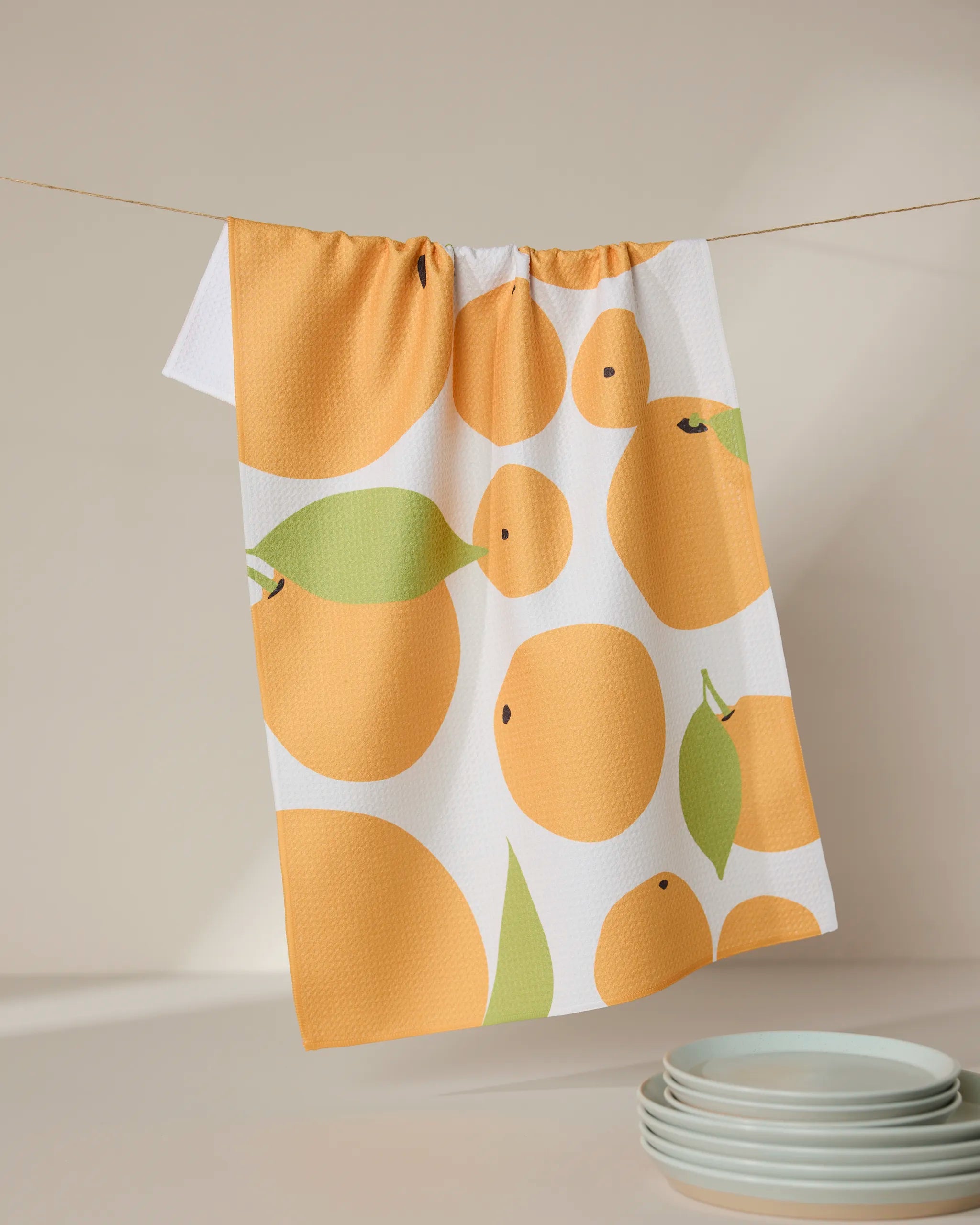 The GEOMETRY Sunny Oranges white kitchen tea towel, crafted from recycled materials, features a bold orange and green fruit print and hangs above stacked white plates on a neutral surface.